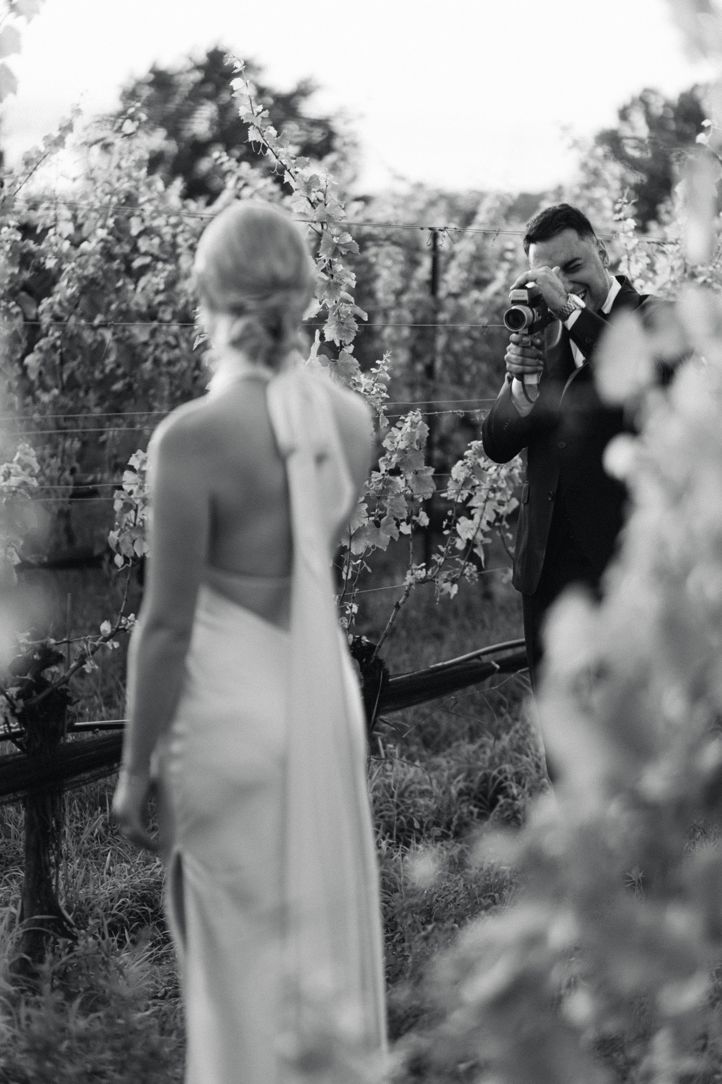 A groom in a vineyard setting taking film of his new bride.