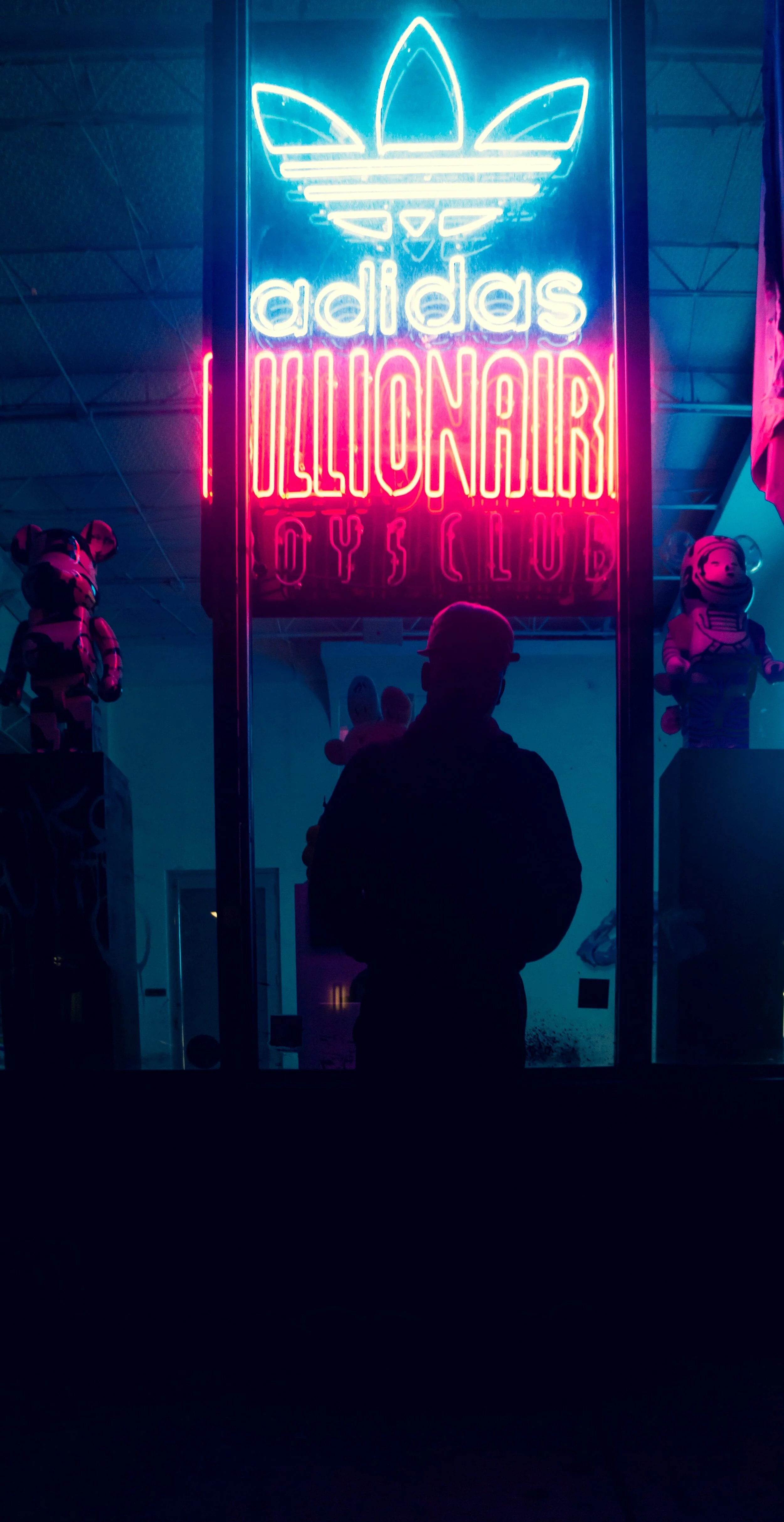 Bright neon sign displaying the Adidas logo and the words 'LIONAIRI' and 'OYS CLUB' at an indoor location. A silhouetted person stands in front of the sign, with toy figures on display behind.