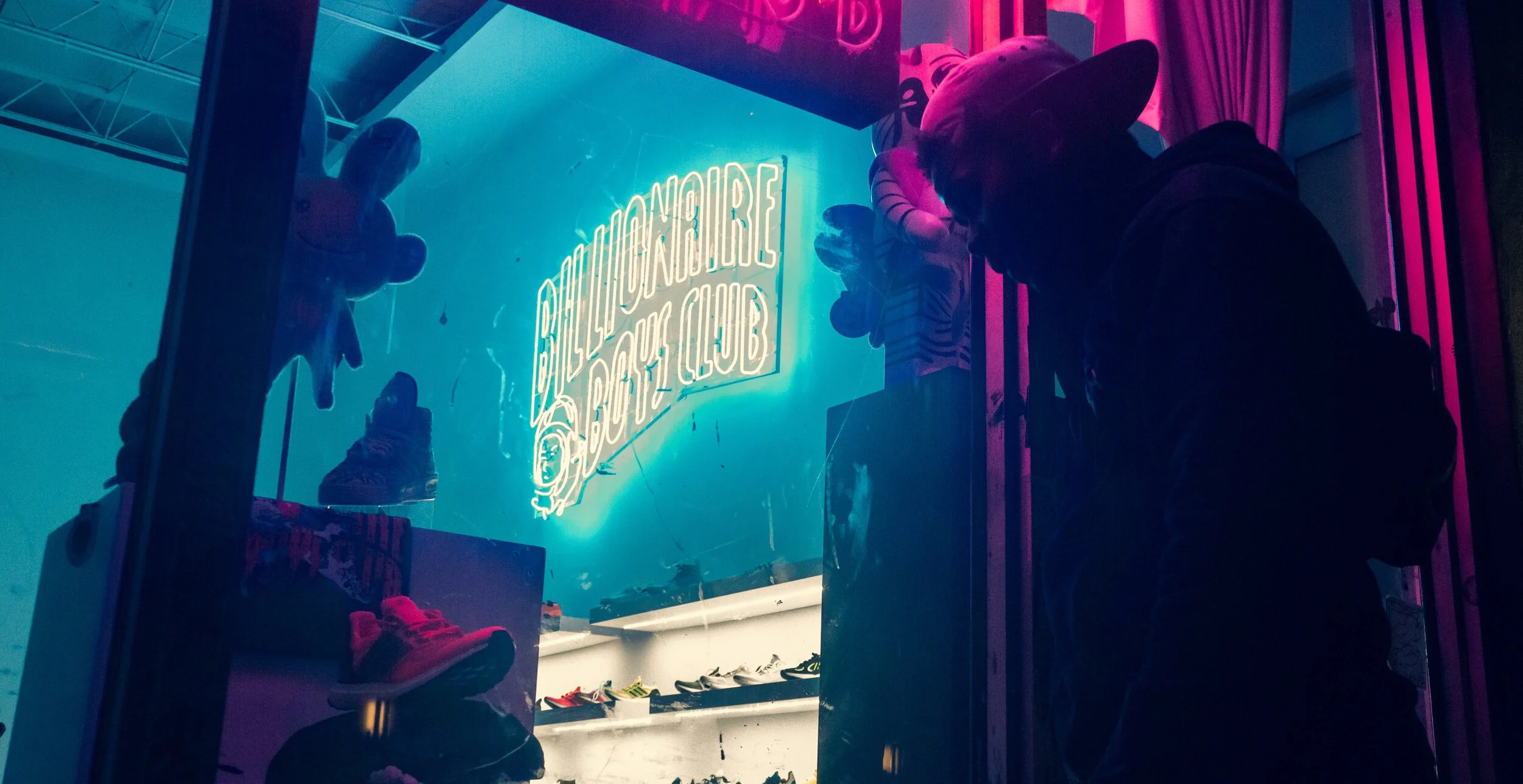 People look into a sneaker store window at night, illuminated by pink and blue neon lights. The store sign reads 'Balenciaga Sport Club' and showcases various sneakers inside.
