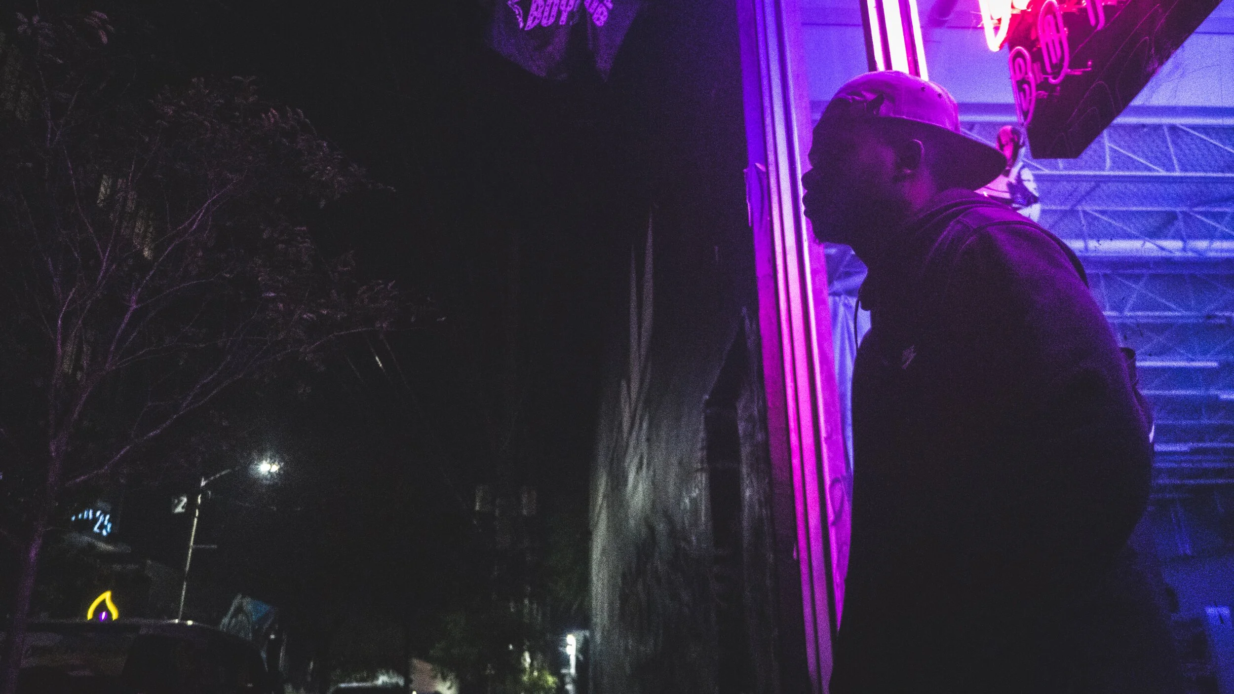 A person standing outside at night near a building with purple neon lights. The person is wearing a cap and a jacket, and is looking at the neon sign.