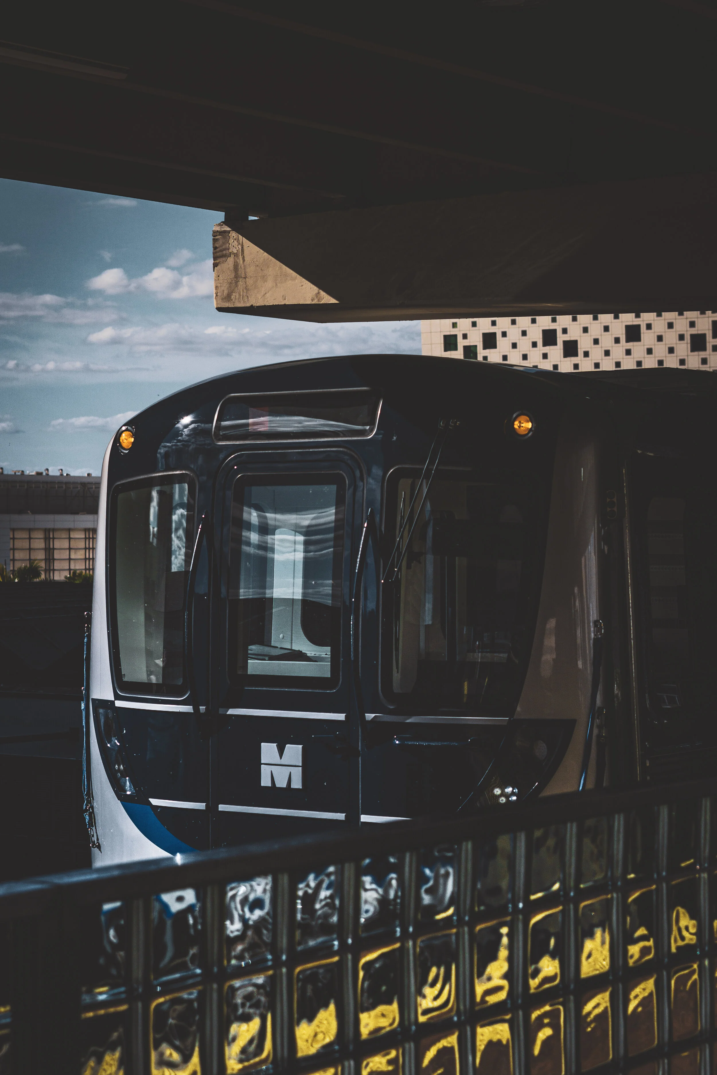 Modern city train station with a black and white train ready for boarding, visible under a concrete overpass, with a cityscape and partly cloudy sky in the background.