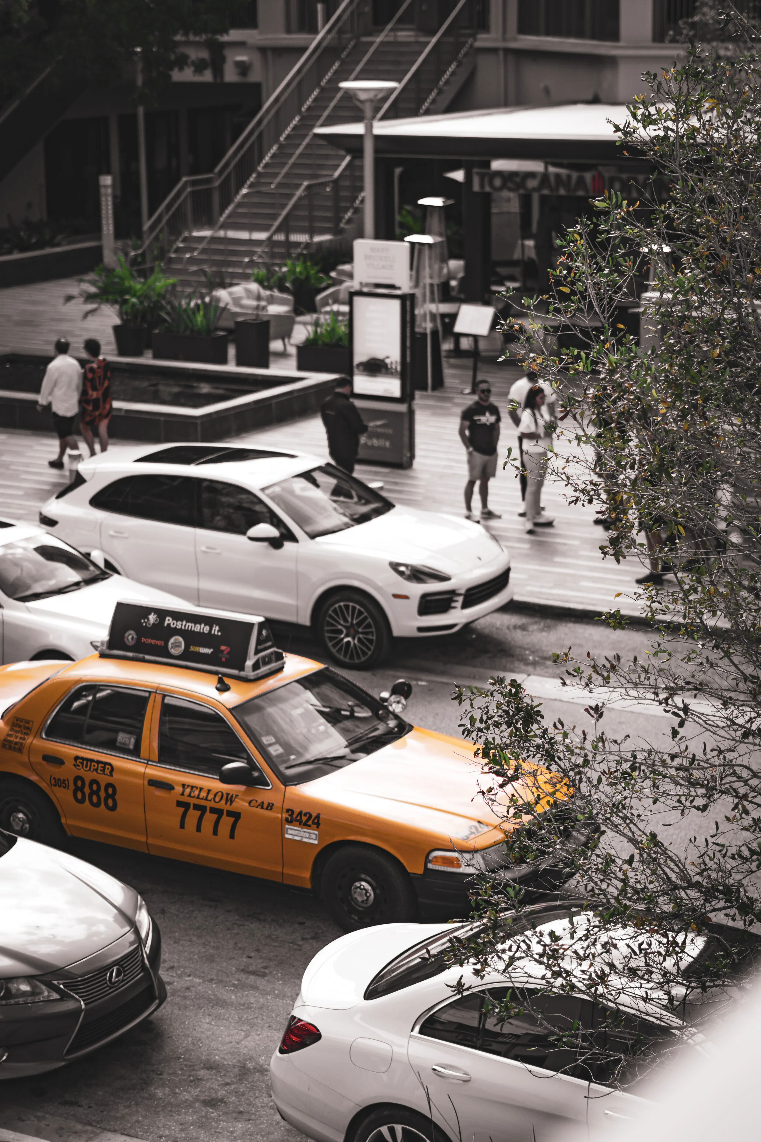Street scene with parked cars, including a yellow taxi, and pedestrians walking near a building with outdoor seating and potted plants.