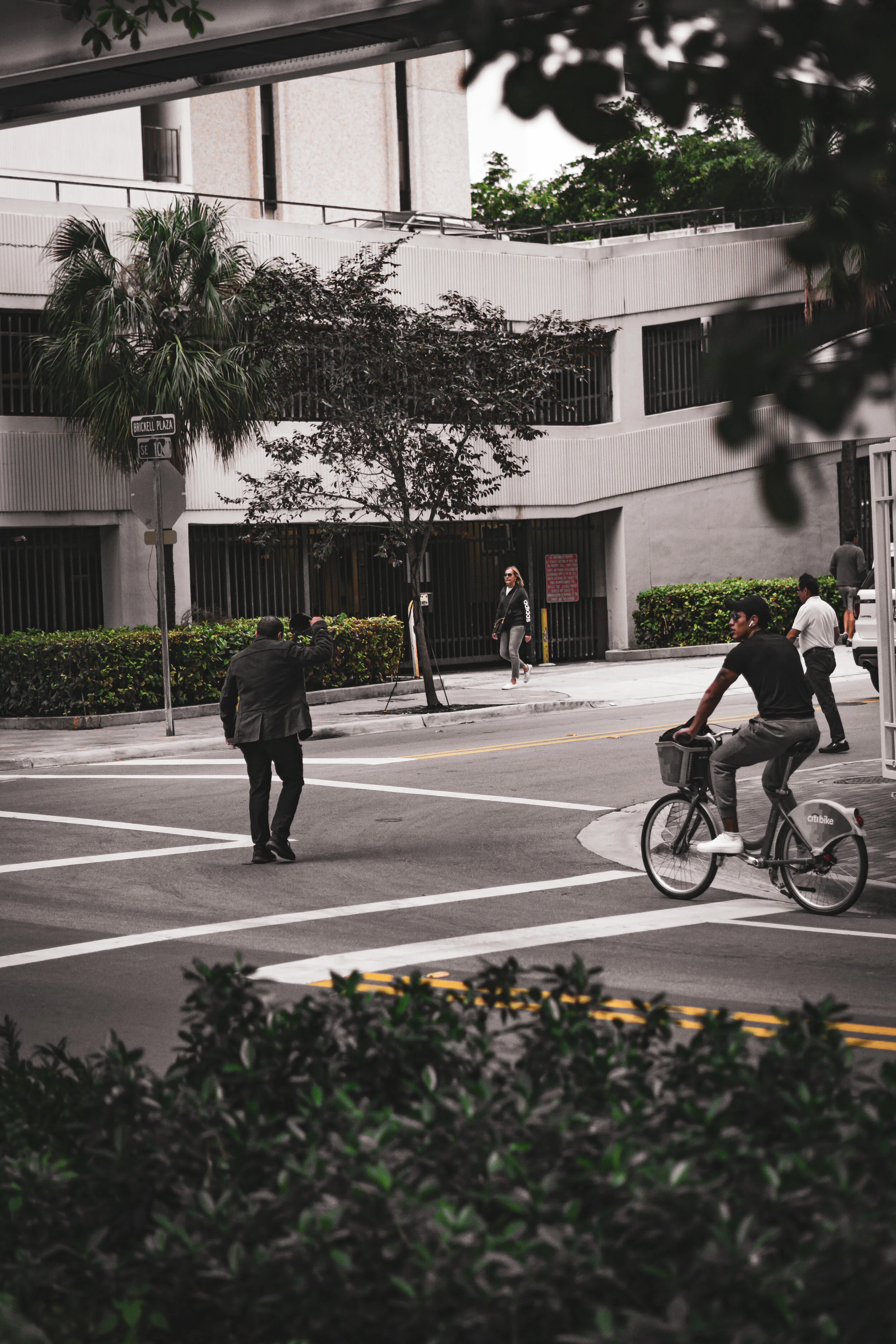 A city street scene with a cyclist crossing a crosswalk, pedestrians walking on the sidewalk, and a modern building in the background, with trees and greenery visible.