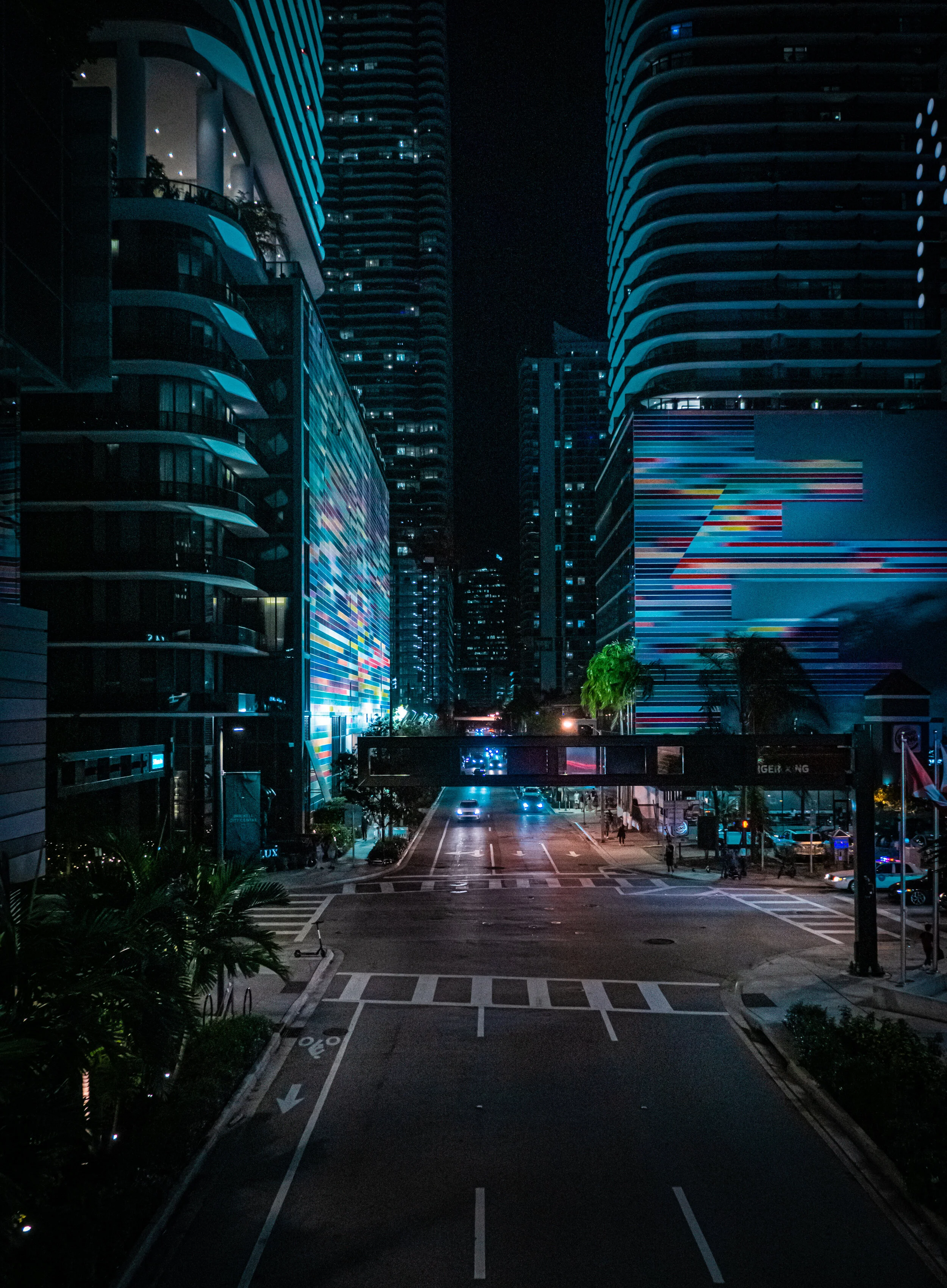 Nighttime city street with tall modern buildings and colorful digital billboards, lightly trafficked with a few cars and pedestrians.