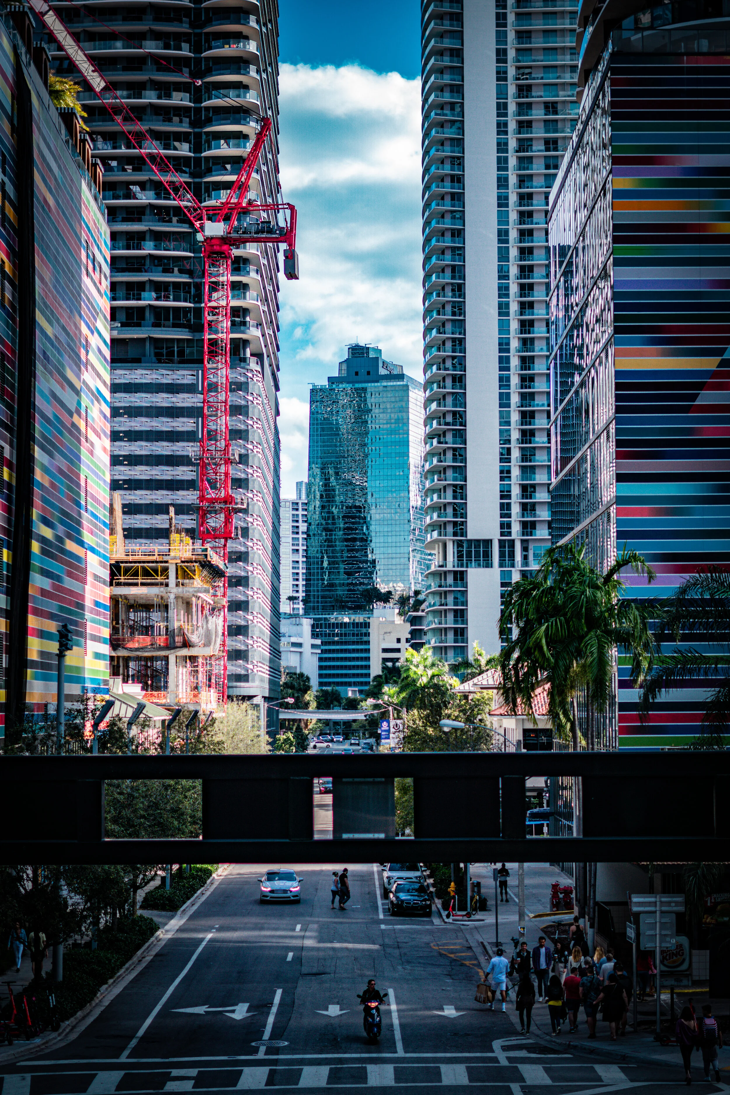 City street scene with tall office buildings, a construction crane, pedestrians, cars, and a motorcycle on a sunny day.