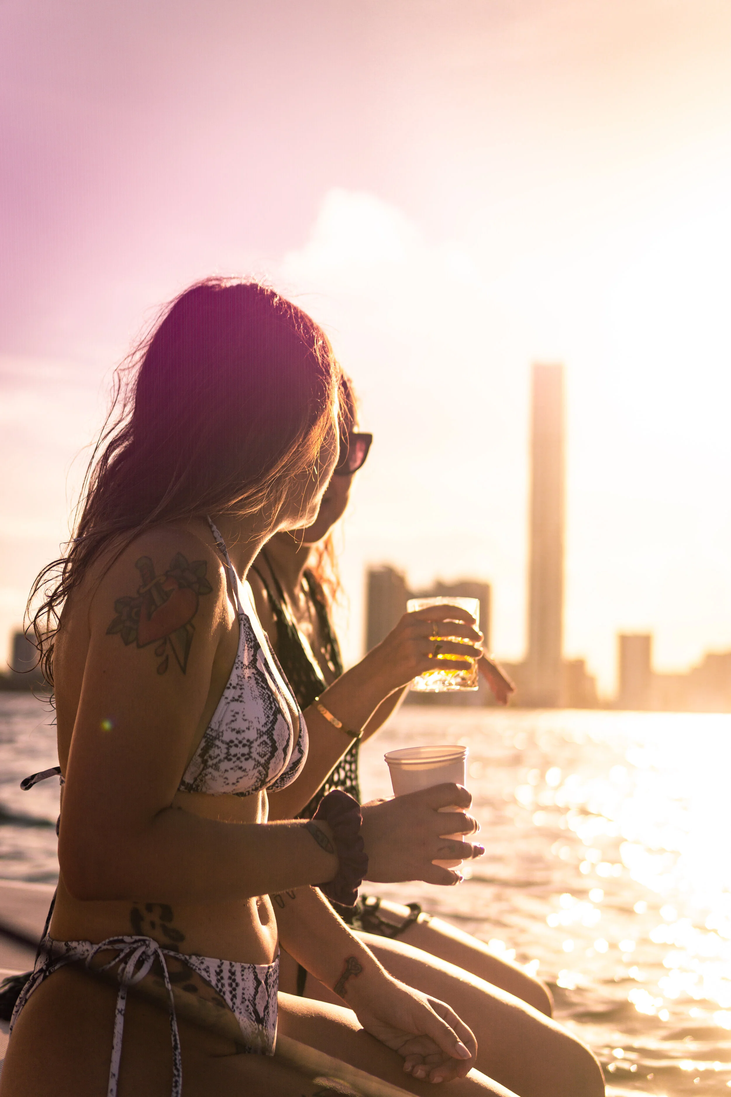 Two women in swimsuits enjoying drinks on a boat during sunset with city skyline in the background.