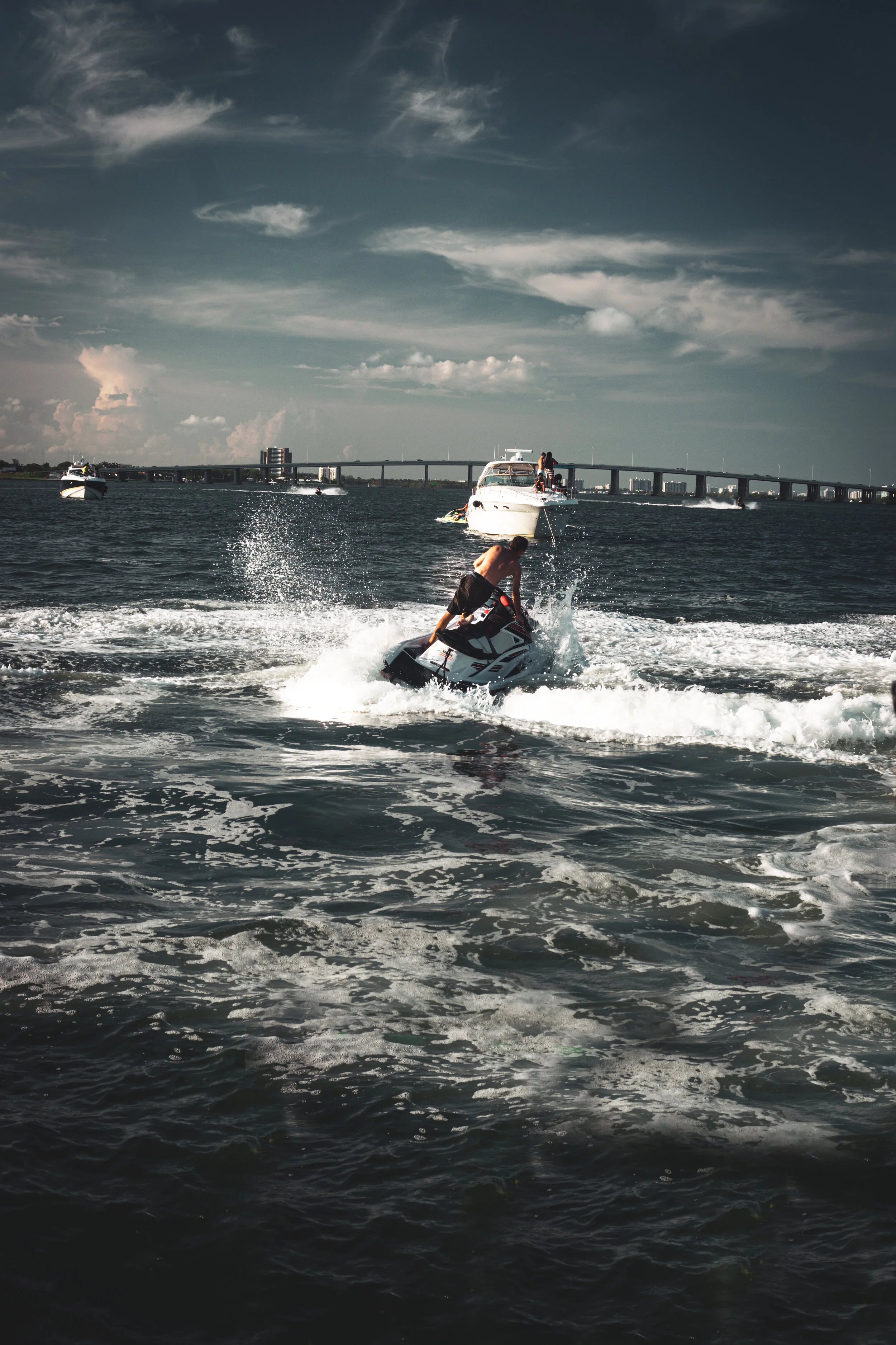 A man riding a jet ski on the water with boats and a bridge in the background under a cloudy sky.