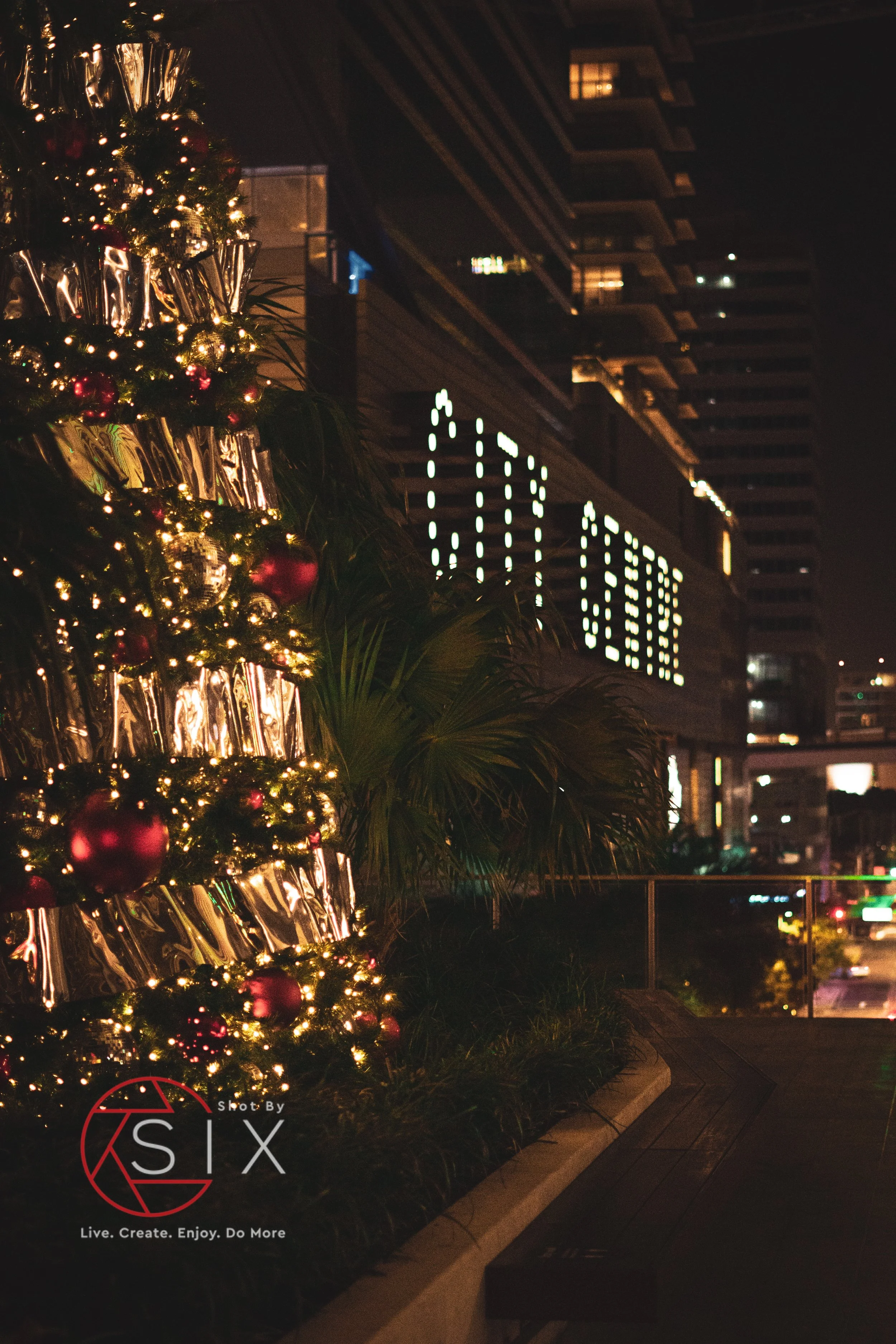 Nighttime cityscape featuring a decorated Christmas tree with lights, ornaments, and metallic ribbons on an outdoor balcony or terrace, with illuminated buildings in the background.