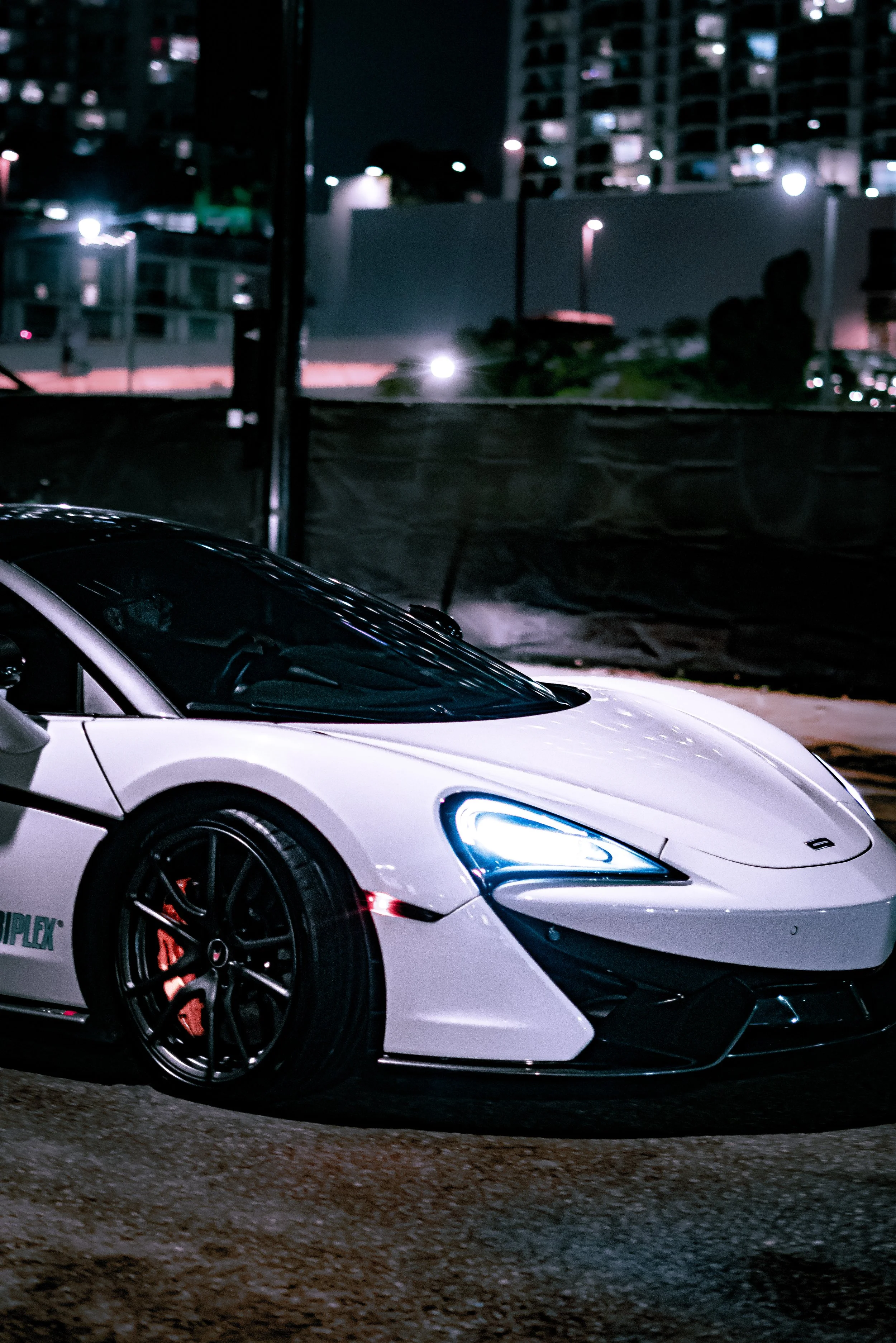 A white race car with black wheels and red brake calipers parked on a city street at night, illuminated by streetlights and city lights in the background.