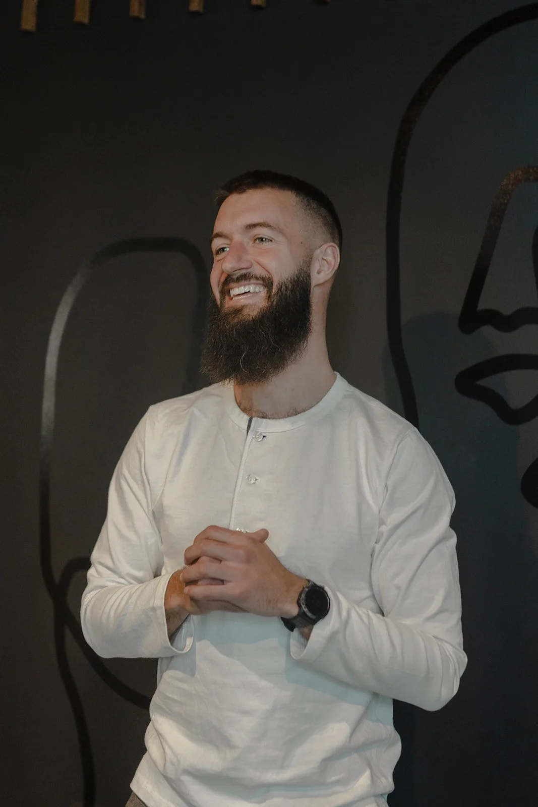 Smiling man with a beard, wearing a white shirt and a watch, standing with hands clasped in front of a dark wall with abstract line art.