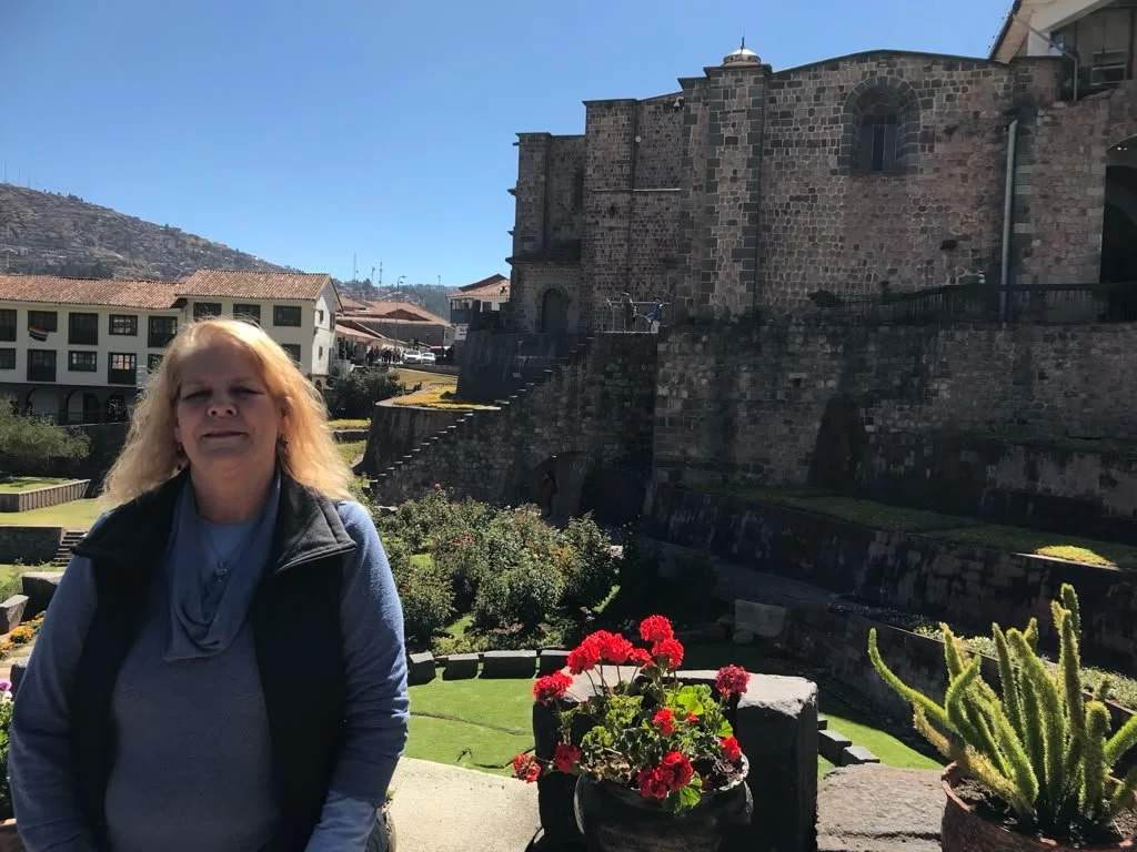 Author and shamanic healer Mona Rain sits in a beam of sunlight illuminating her light blonde hair and soft blue sweater in the gardens of the Qorikancha Temple in Cusco, Peru.