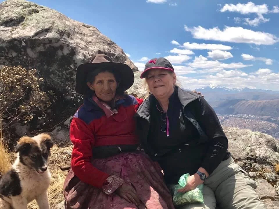 Paqo dona Maria sits with Mona and a dog pose beside a large rock on a mountain overlook with the distant Sacred Valley and blue sky of Peru.