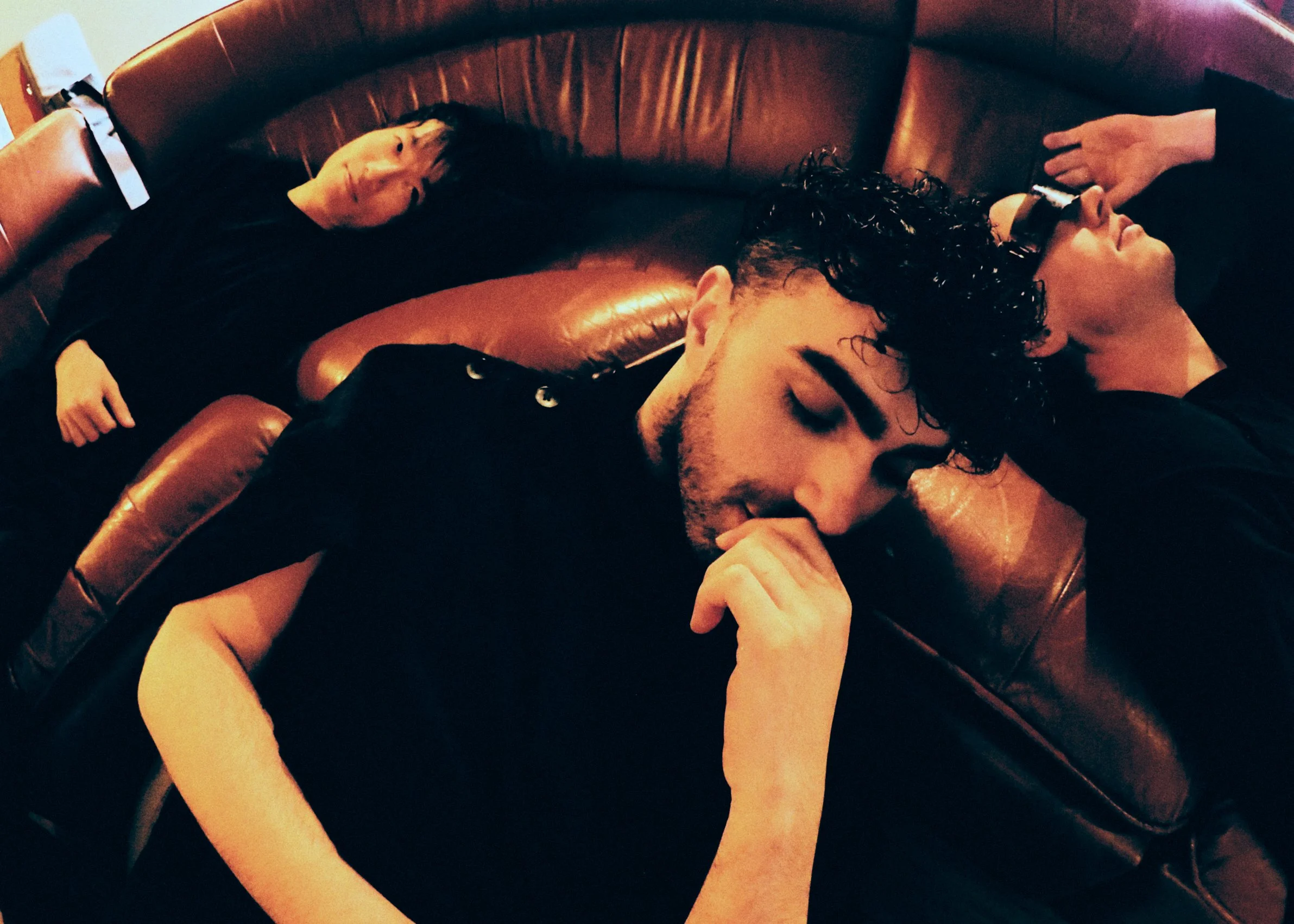 Three people lying on a leather couch, with one person wearing glasses and curly hair, a second person with short dark hair, and a third person with a person with detached arm with a phone.