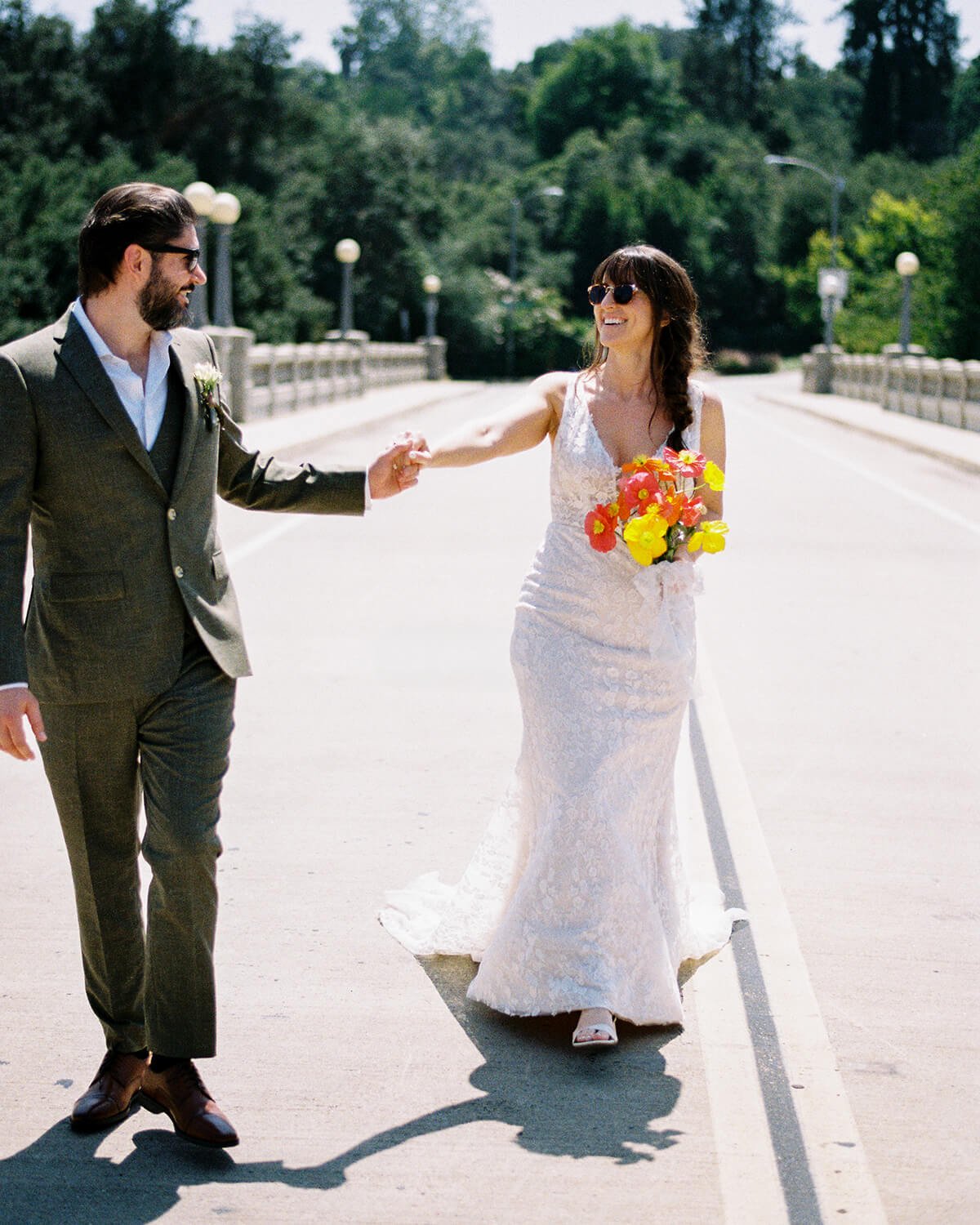 A bride and groom holding hands and smiling on a sunny day in the middle of an empty road surrounded by trees.