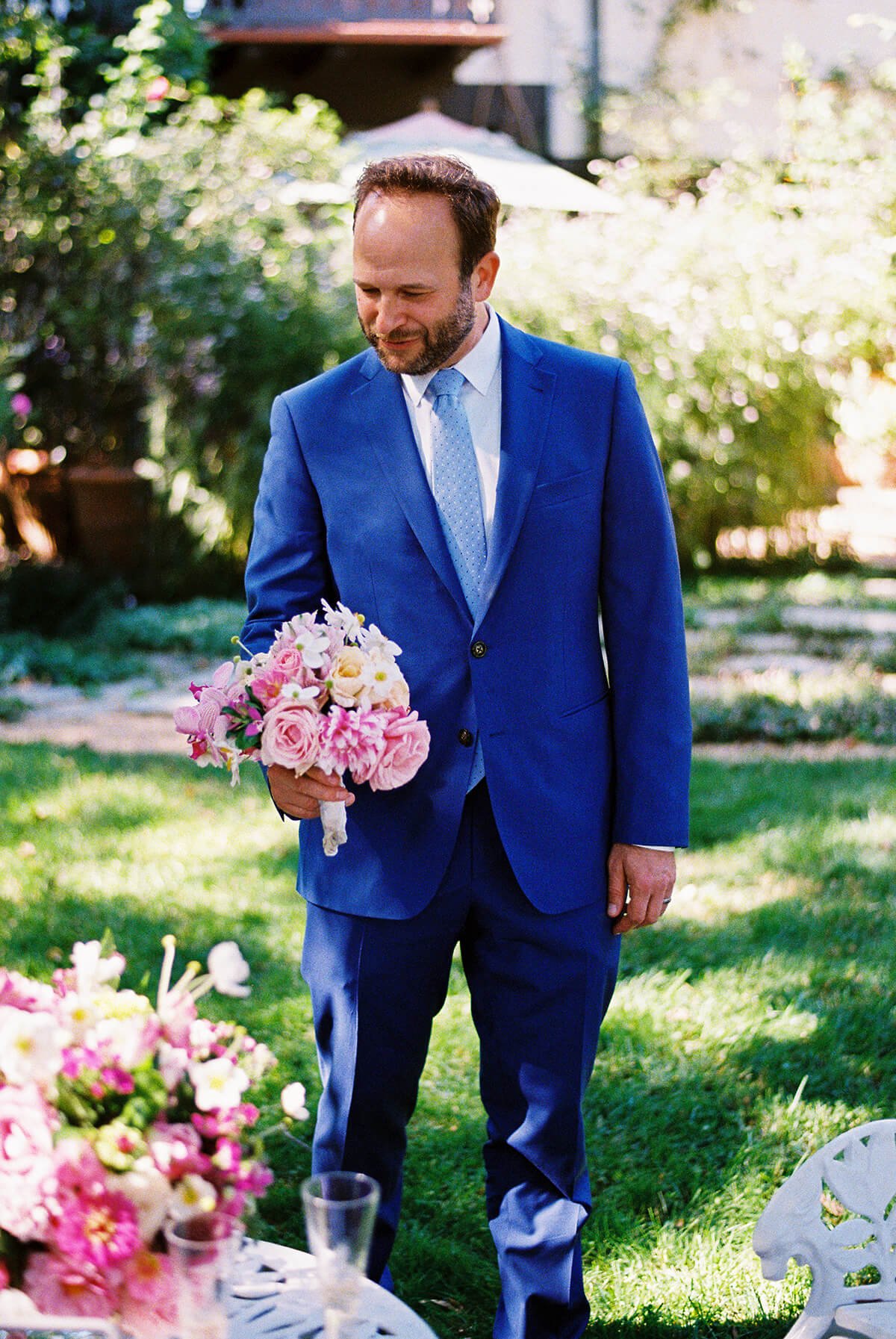 A man in a blue suit holding a bouquet of pink and white flowers outdoors on a sunny day.