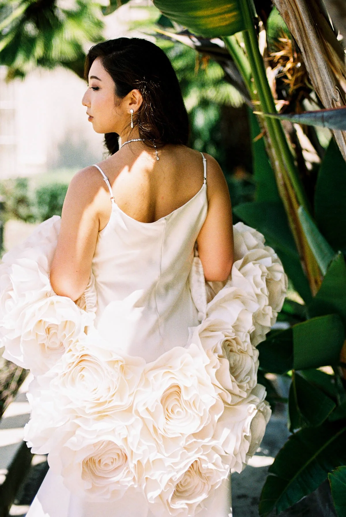 Woman in a white dress with large floral ruffle details, standing among green tropical plants, turned sideways with her eyes closed.