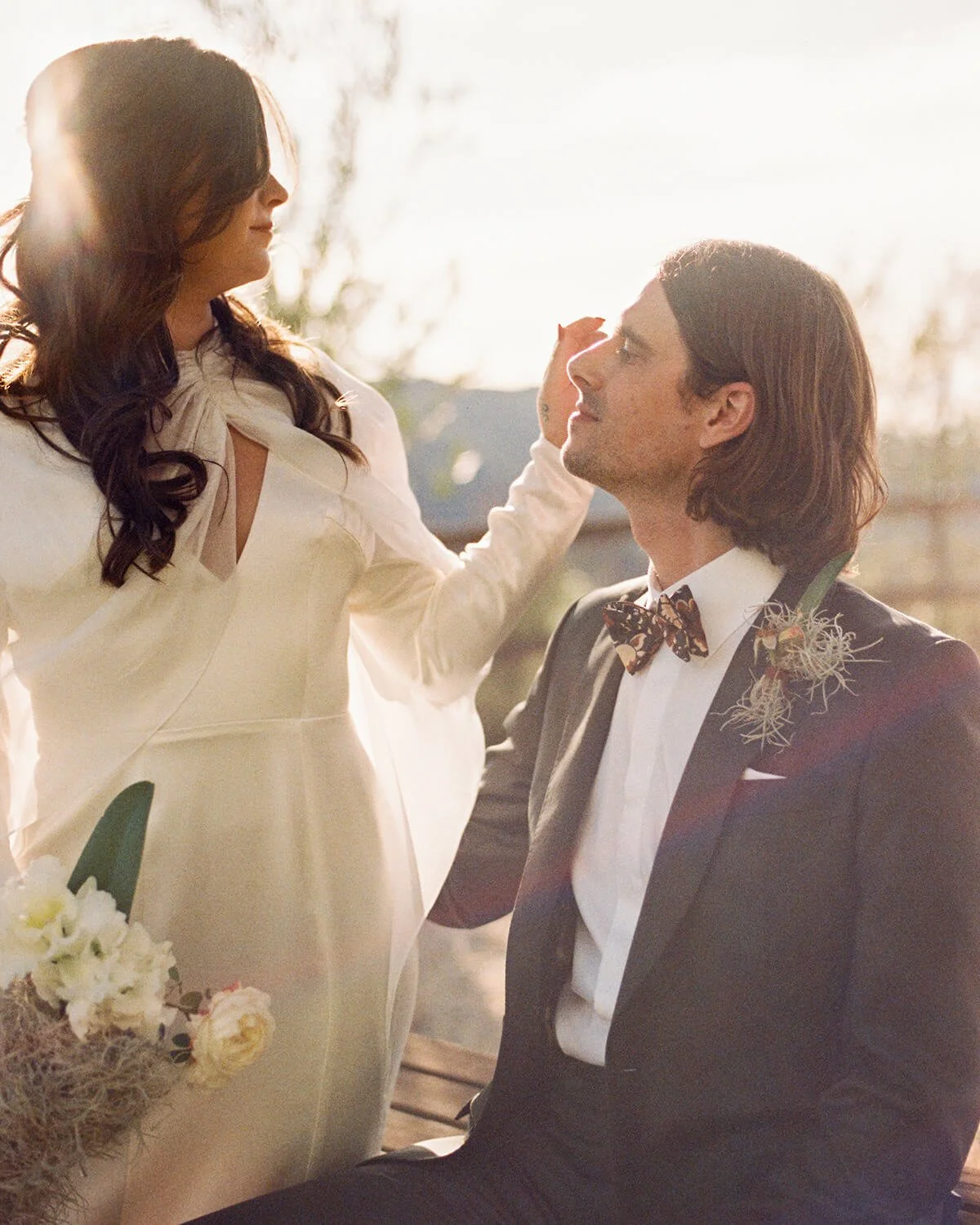 A woman in a white dress and a man in a black tuxedo with a bow tie sit outdoors during sunset, looking at each other, with the woman gently touching the man's face.