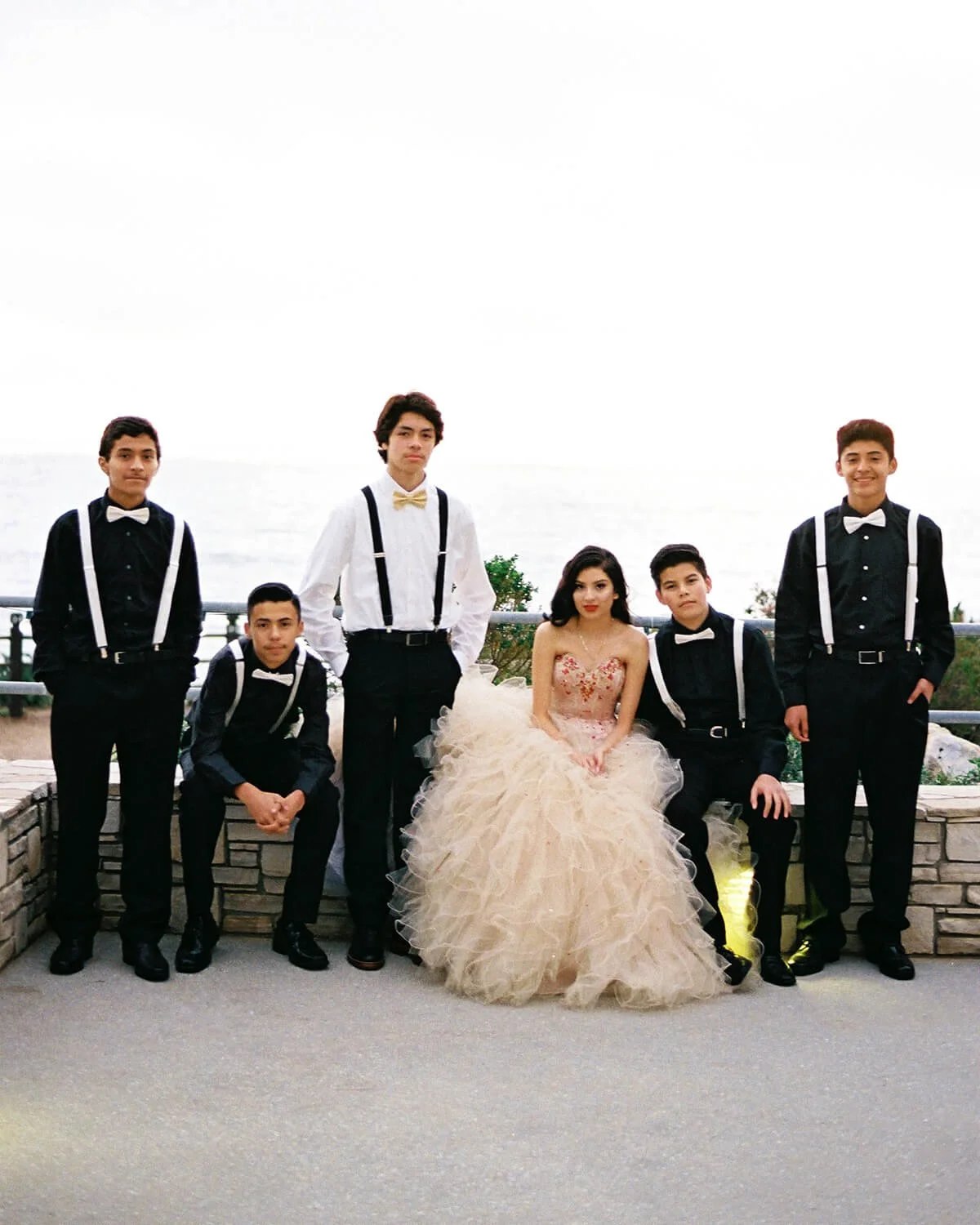 A Quinceañera in a fancy gold dress with six chambelanes in formal wear posing outdoors by a stone wall, with a cloudy sky and ocean in the background.