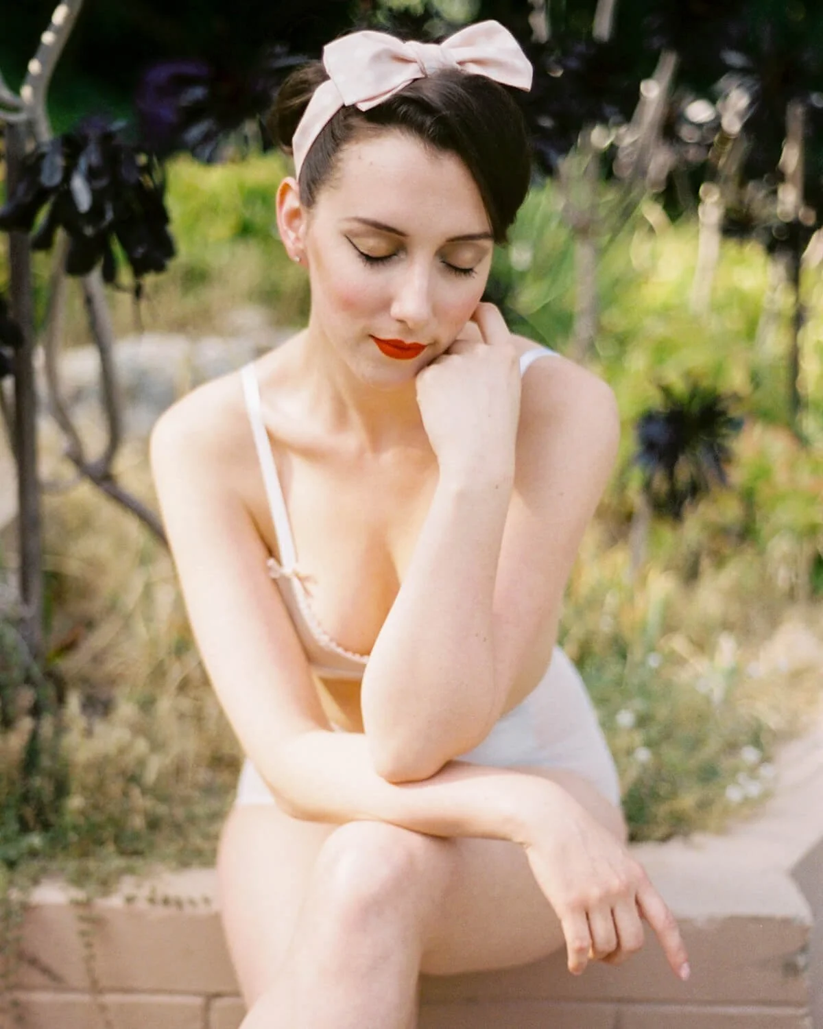 A woman with short dark hair, wearing a pink bow headband and a light-colored bra, sitting outdoors with her eyes closed, resting her chin on her hand, and smiling softly.