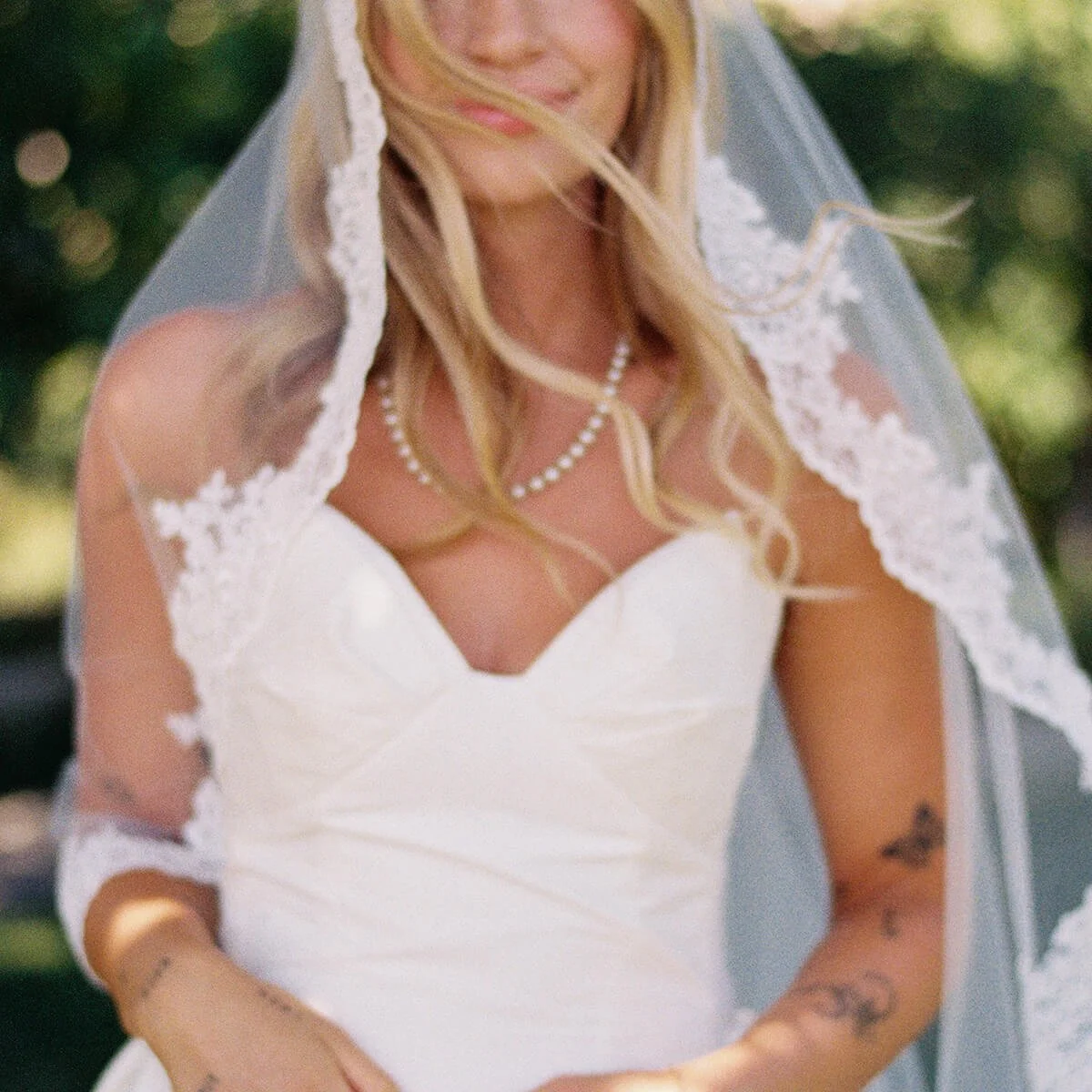 A woman wearing a white wedding dress with lace details and a veil, smiling outdoors.