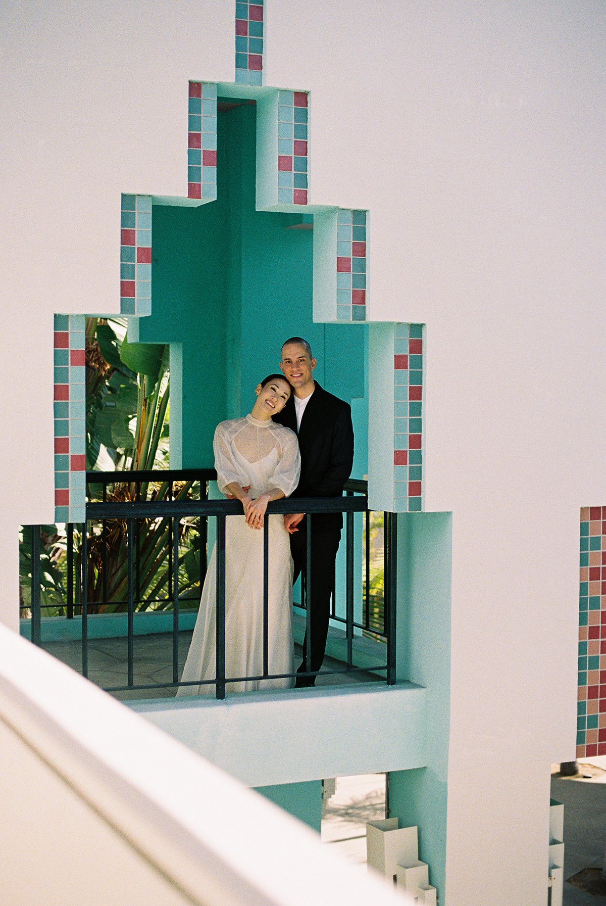 A couple dressed in wedding attire standing on a balcony with a turquoise and pink tiled architectural backdrop, smiling at the camera.