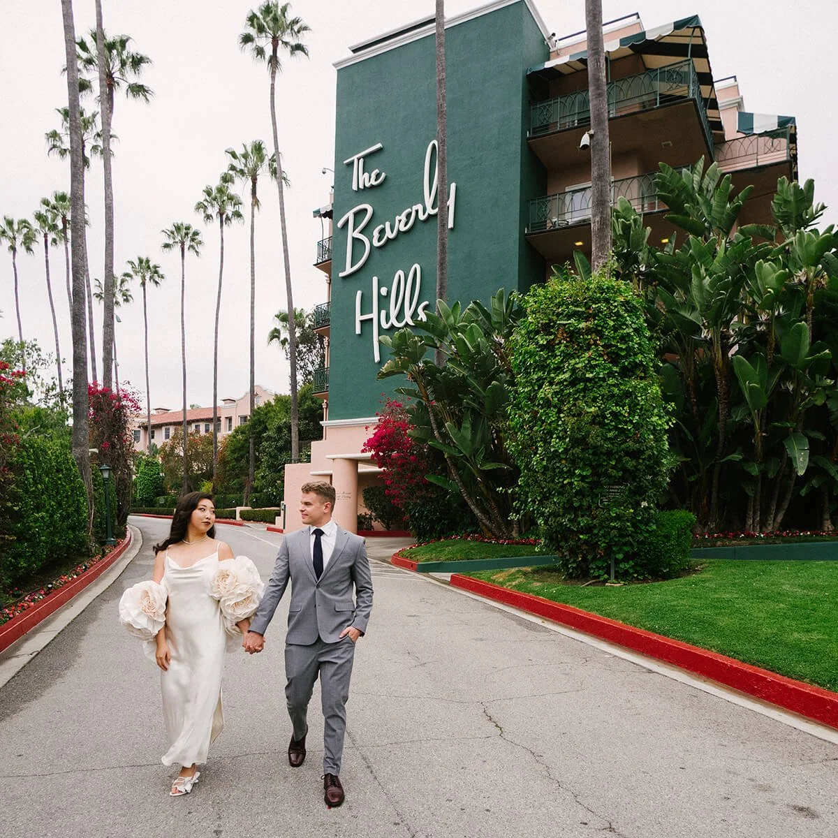 A couple dressed in wedding attire walking hand in hand in front of The Beverly Hills Hotel in an overcast morning, surrounded by palm trees and lush greenery.