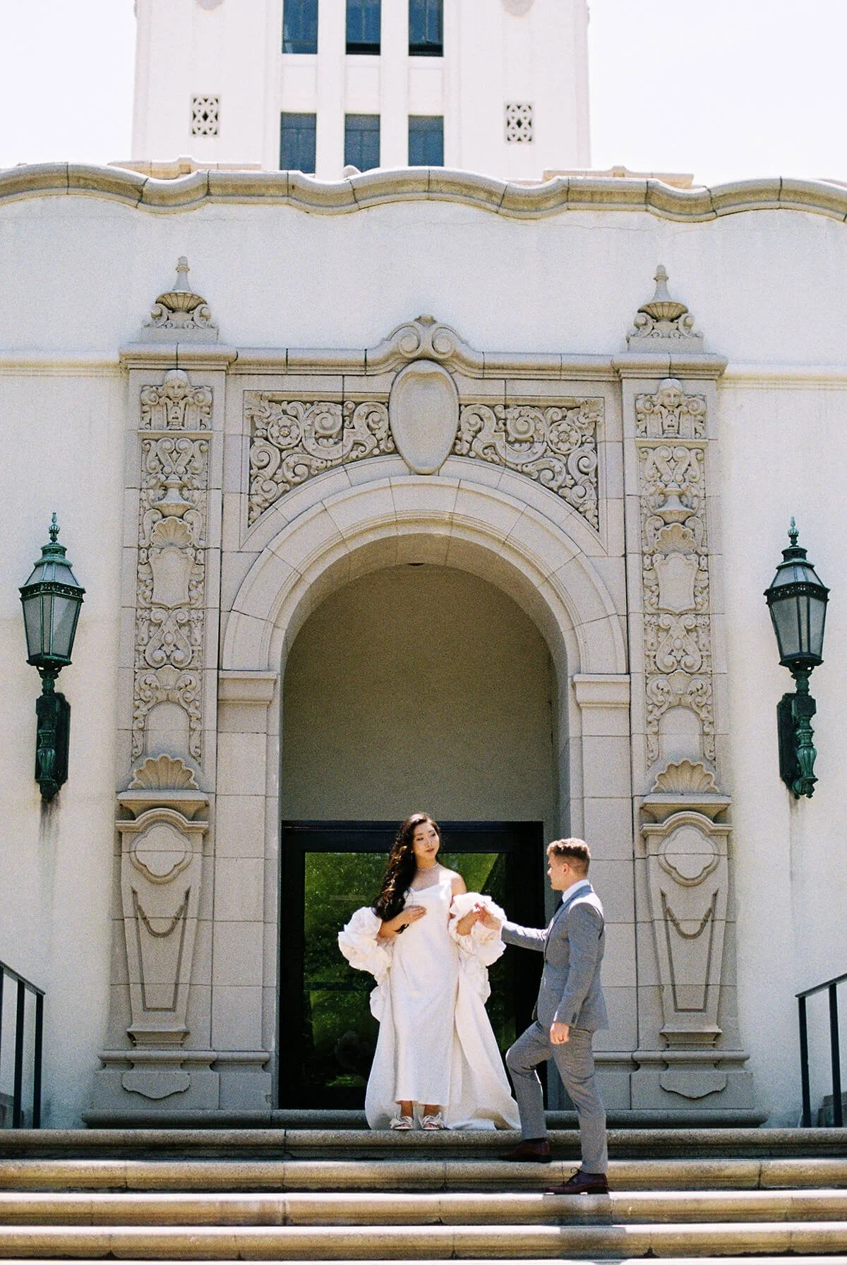  Bride and groom pause on the grand staircase outside Beverly Hills City Hall, framed by carved stone columns and archways, just after their Beverly Hills courthouse marriage ceremony. 