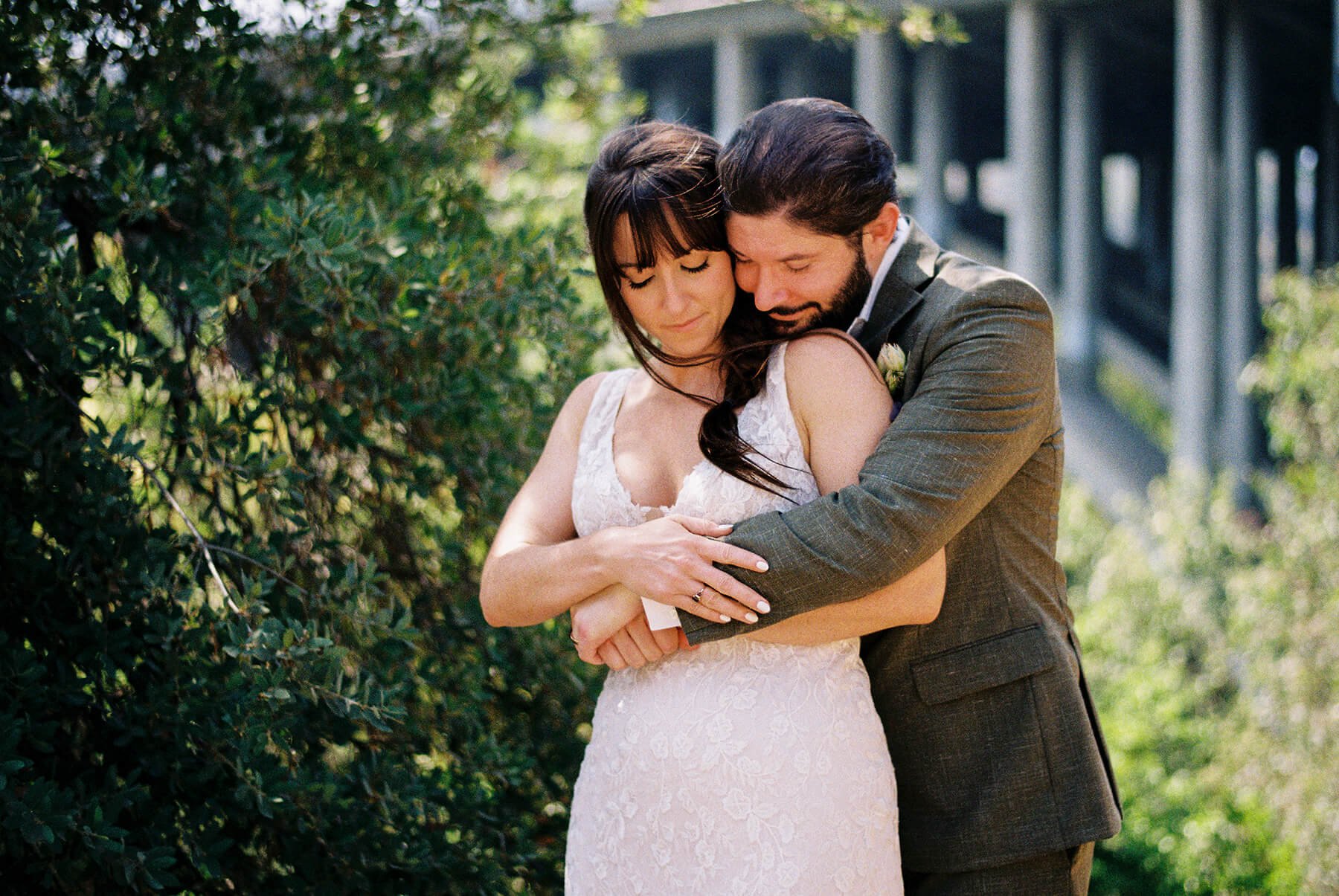 A bride and groom embracing outdoors, with the bride in a white lace wedding dress and the groom in a gray suit, surrounded by greenery.