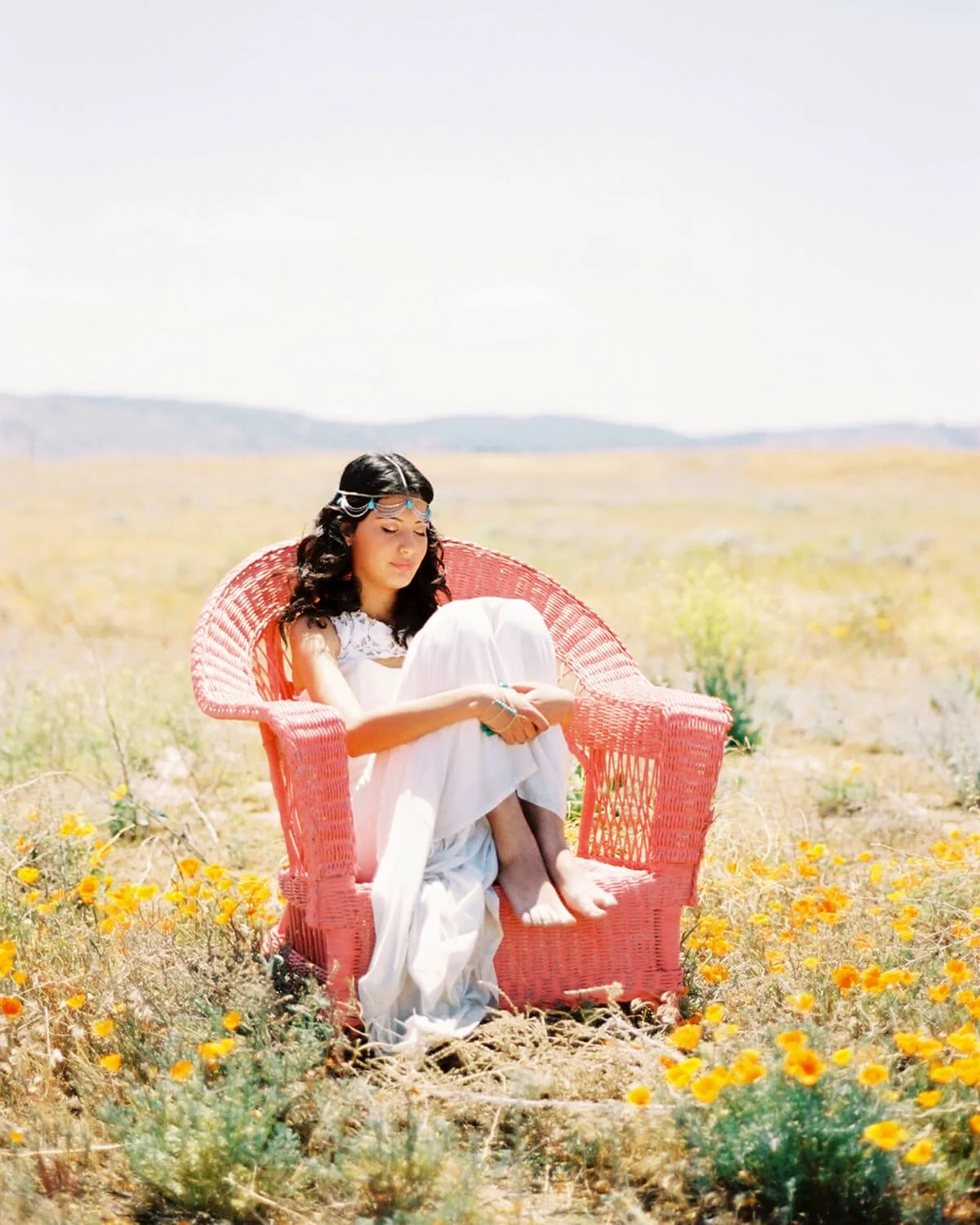 A Quinceañera sitting on a pink wicker chair in a field of yellow flowers on a sunny day, wearing a white dress and sunglasses, with her eyes closed.