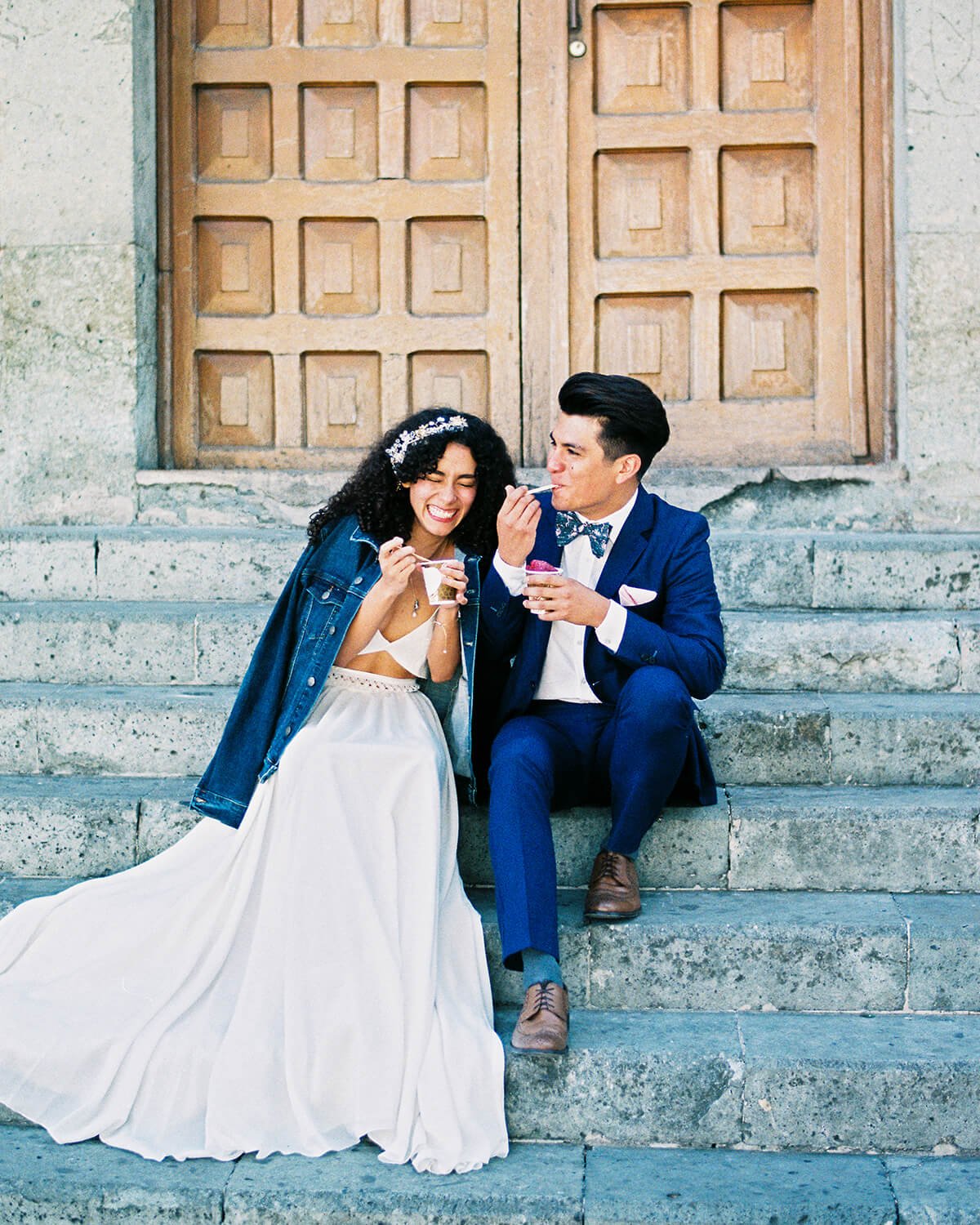A bride and groom sitting on stone steps in front of a wooden door, sharing ice cream and laughing.