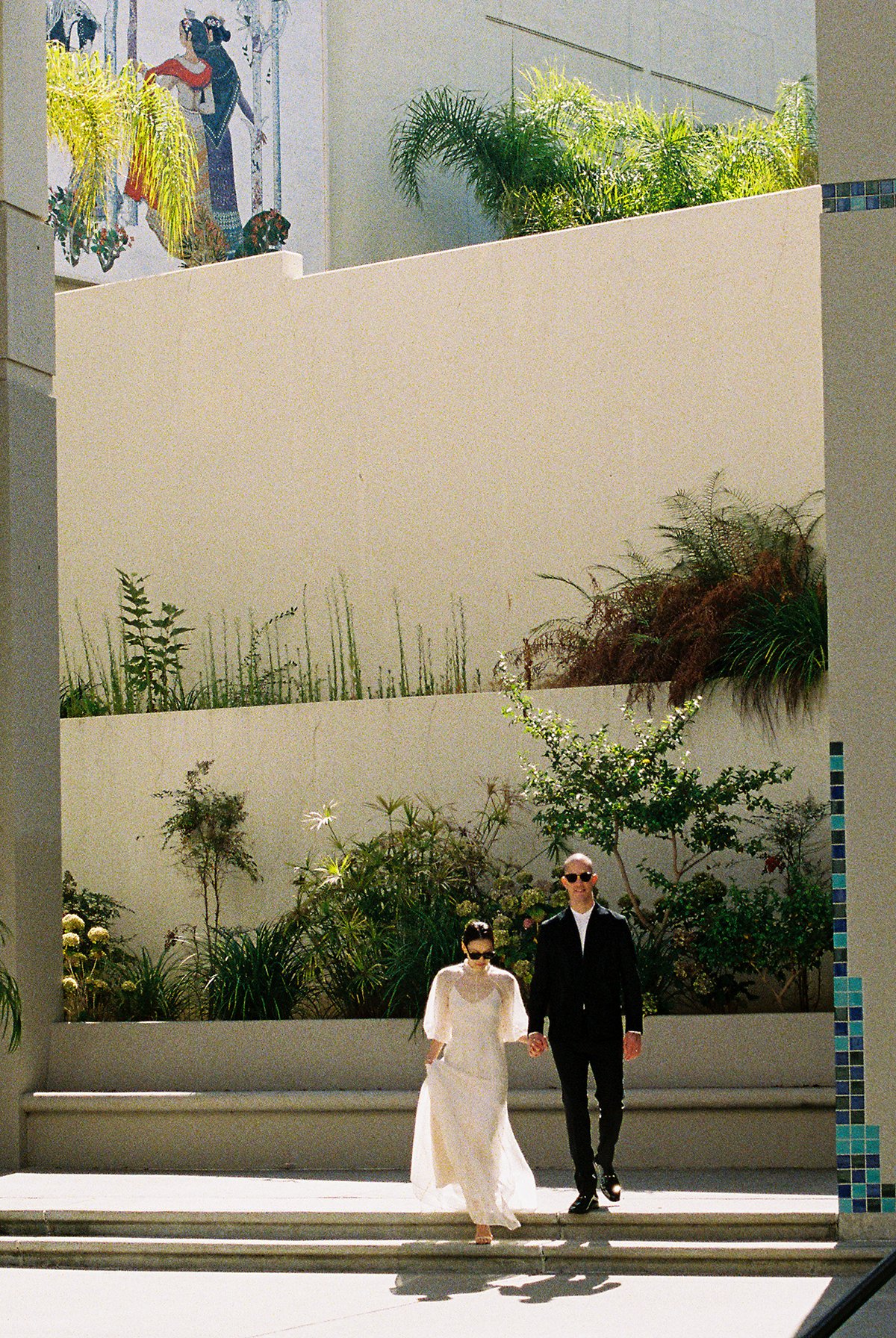 A couple dressed in wedding attire walking hand in hand outdoors on a sunny day, with a modern landscaped background featuring plants, a white wall, and colorful artwork.