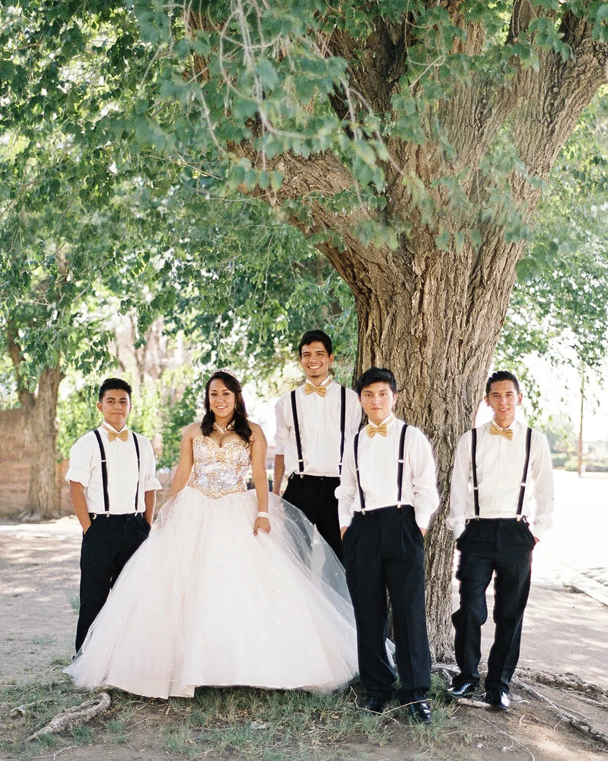 A Quinceañera in a white gown with lace details standing under a tree, surrounded by four chambelanes in white shirts, black suspenders, and gold bow ties, outdoors on a sunny day.