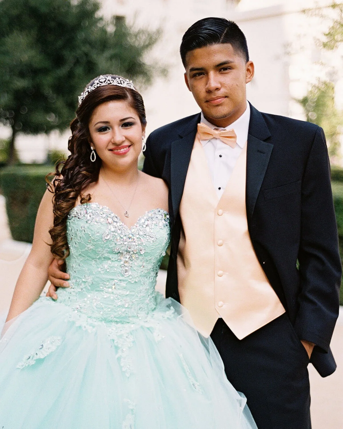 A Quinceañera wearing a light-colored wedding gown with detailed beadwork and a tiara, and the chambelan in a black tuxedo with a beige vest and bow tie.