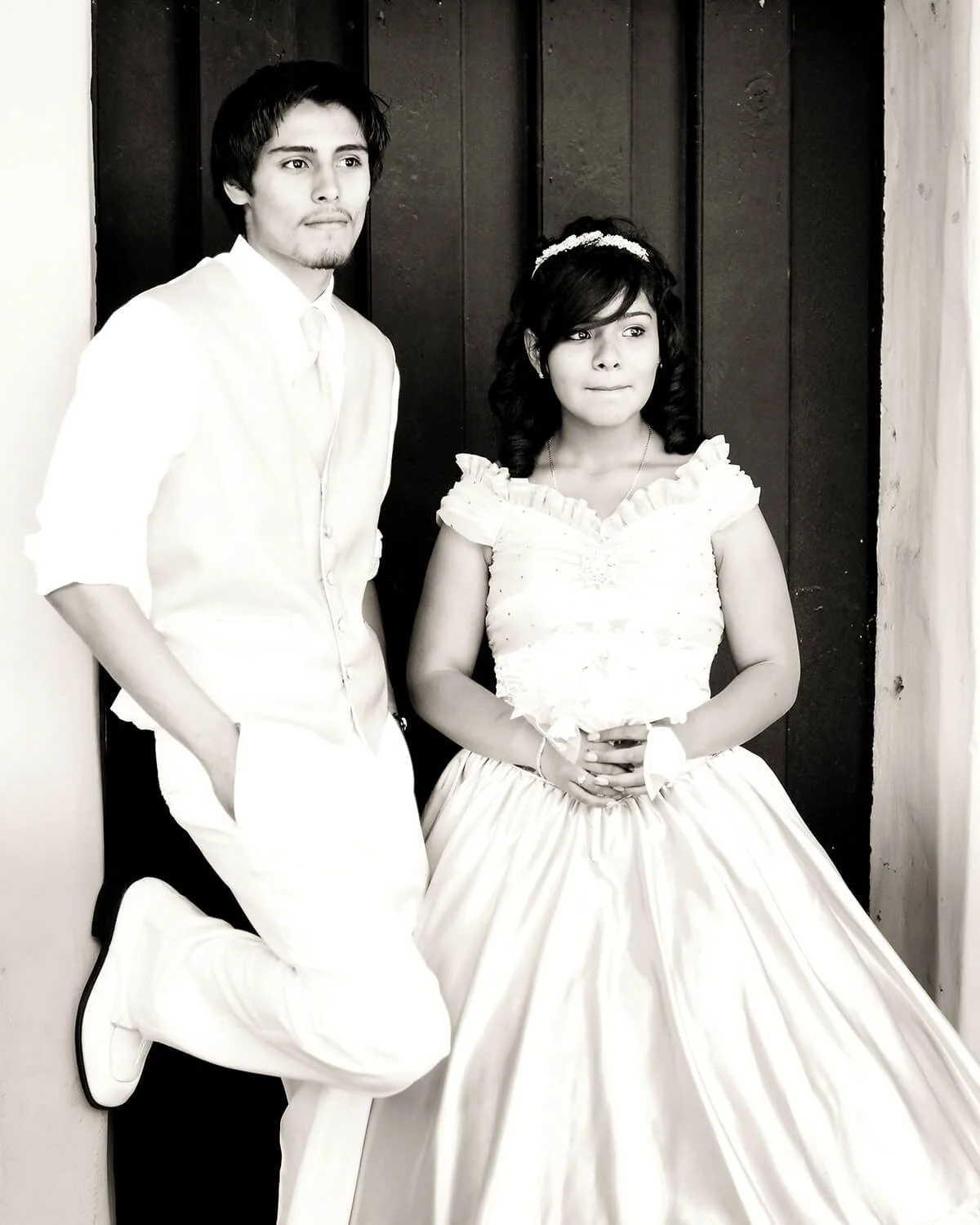 A black and white photo of a Quinceañera and chambelan de honor standing in front of a dark wooden wall, dressed in formal attire, with the woman wearing a wedding gown.