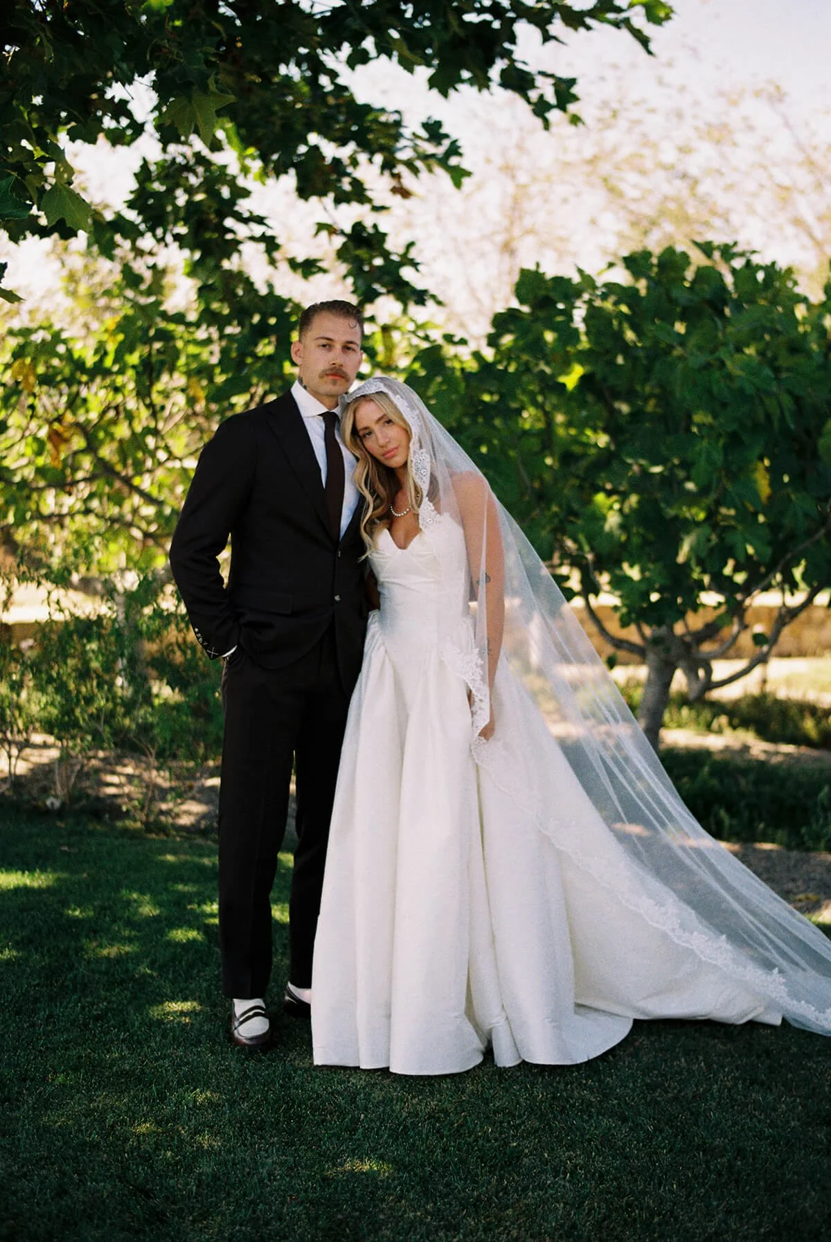 A bride and groom standing outdoors under trees. The groom is wearing a black suit with a white shirt and tie. The bride is in a white wedding dress with a veil and has her head resting on the groom's shoulder. They are on a grassy area with lush gre