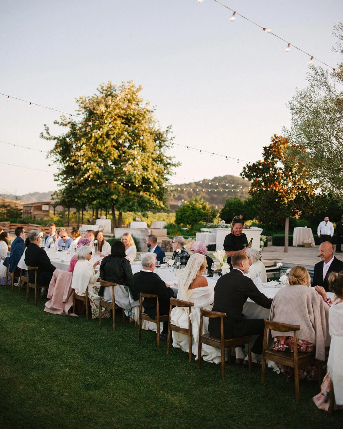 People seated at a long outdoor banquet table during a wedding reception at sunset, with string lights overhead and trees in the background.