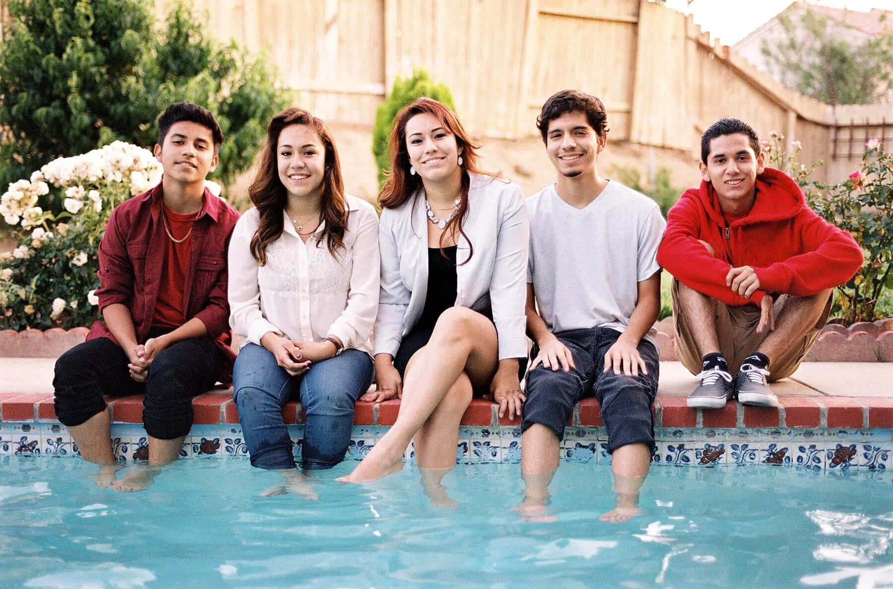 A Quinceañera court sitting on the edge of a swimming pool with their feet in the water, smiling at the camera, in a backyard with a wooden fence and lush greenery.