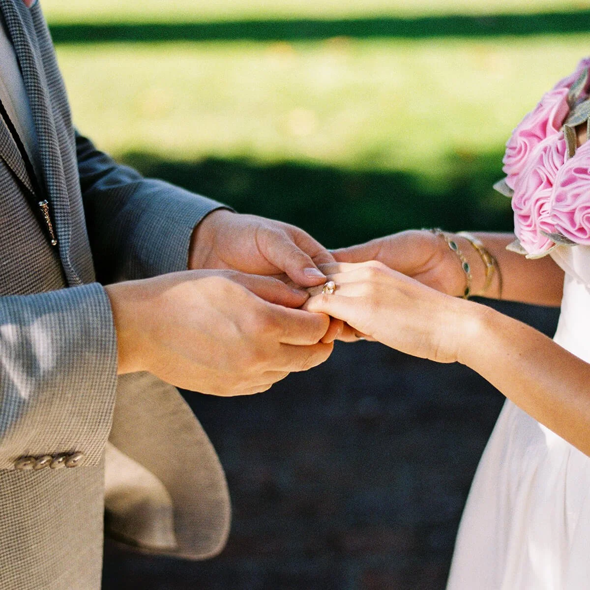 Close-up of a wedding ceremony where a groom is placing a ring on a bride's finger, with a bouquet of pink flowers in the background.