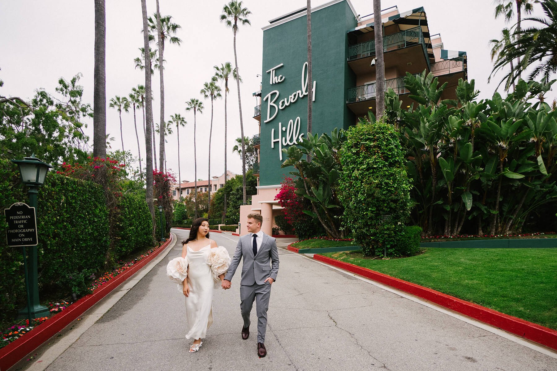 A couple dressed in wedding attire walking hand in hand near The Beverly Hills sign, surrounded by palm trees and lush greenery.