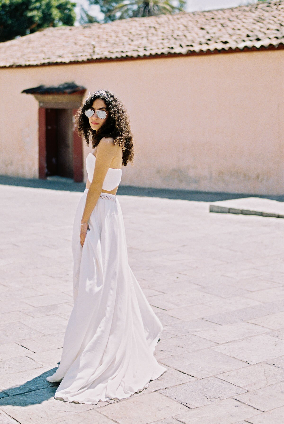 Young woman in a white strapless top and flowing skirt standing outdoors on a sunny day, wearing sunglasses and curly hair, with a pastel-colored wall and blue sky in the background.