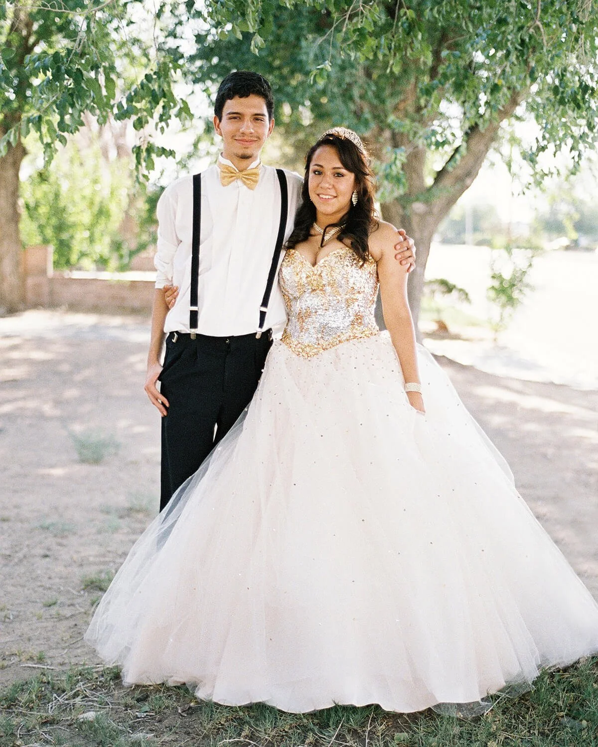 A chambelan wearing a white dress shirt with suspenders, a gold bow tie, and black pants. A Quinceañera wearing a gold and white gown with intricate gold detailing on the bodice and a full tulle skirt. They are smiling and standing close together.