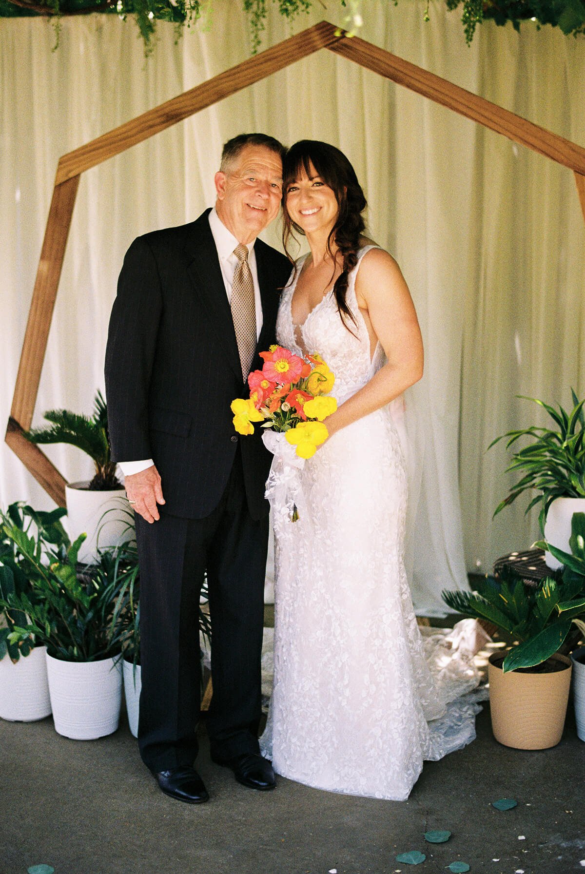 A bride in a white lace wedding dress holding a colorful bouquet, standing next to an older man in a black suit, both smiling, in a decorated wedding setting with potted plants and wooden geometric backdrop.