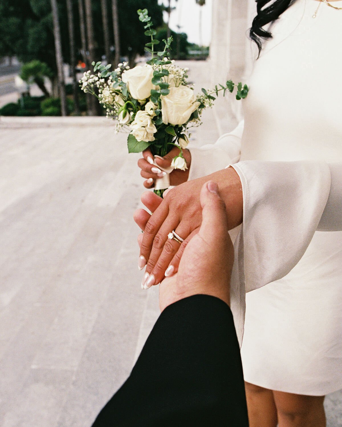 A woman in a white dress is holding a small bouquet of white roses and greenery while a person, dressed in black, is holding her hand showing a wedding ring.