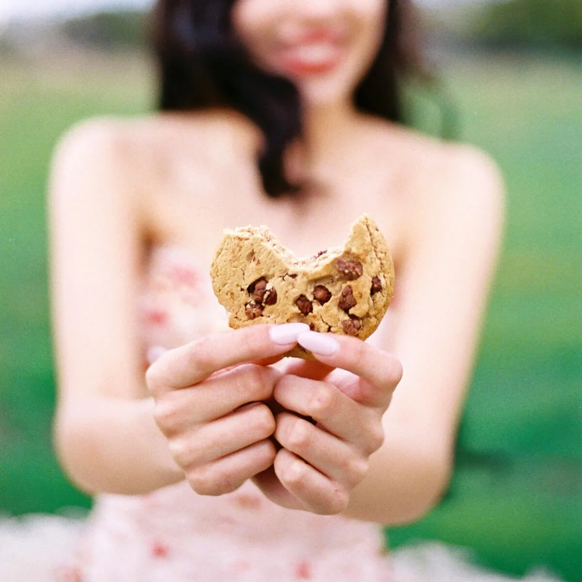 A Quinceañera holding a bitten chocolate chip cookie outdoors with a grassy background.