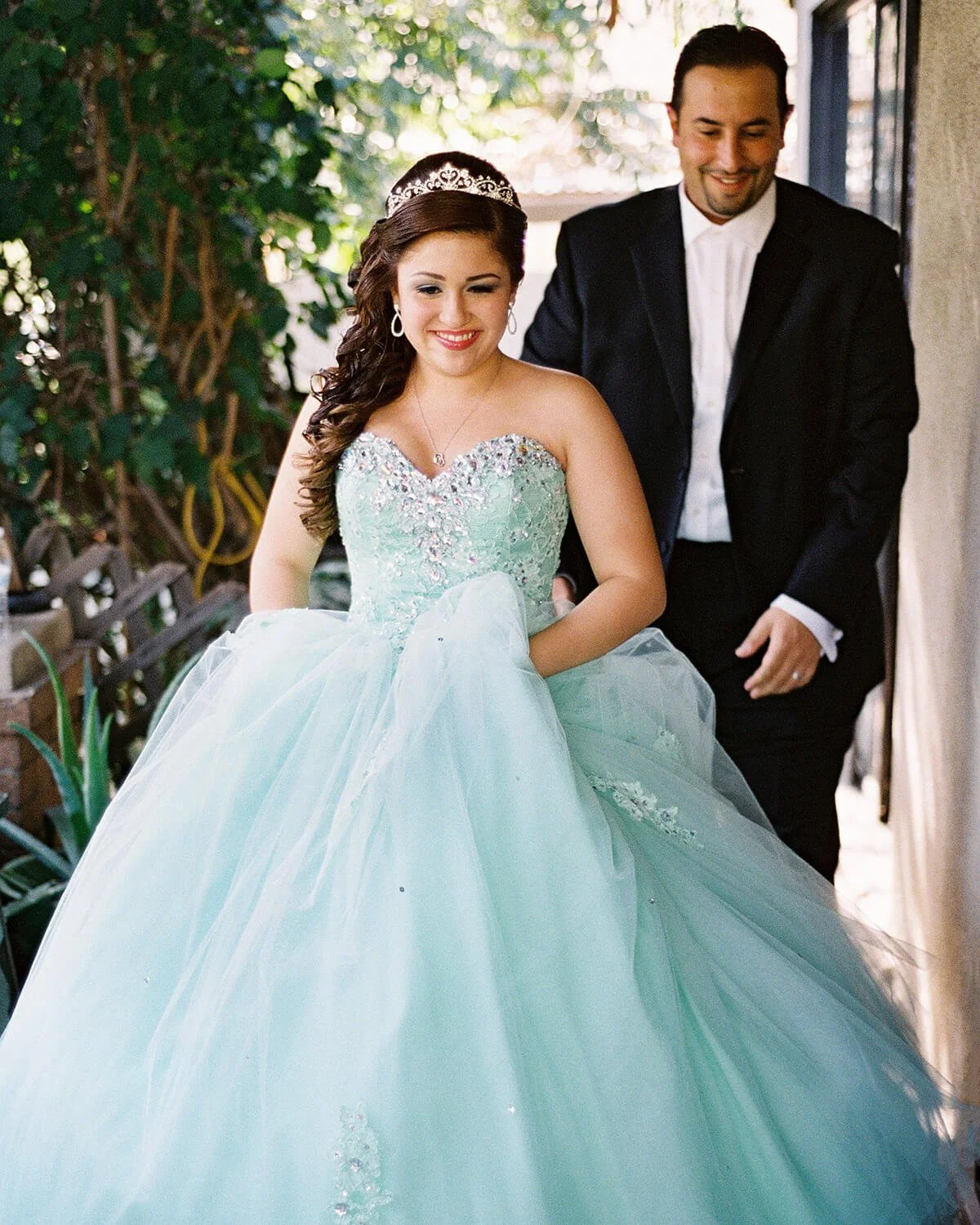 A young woman in a light blue quinceañera dress with a tiara, smiling and holding the skirt of her gown. A man in a black suit with a white shirt stands behind her, also smiling, in an outdoor setting with greenery.