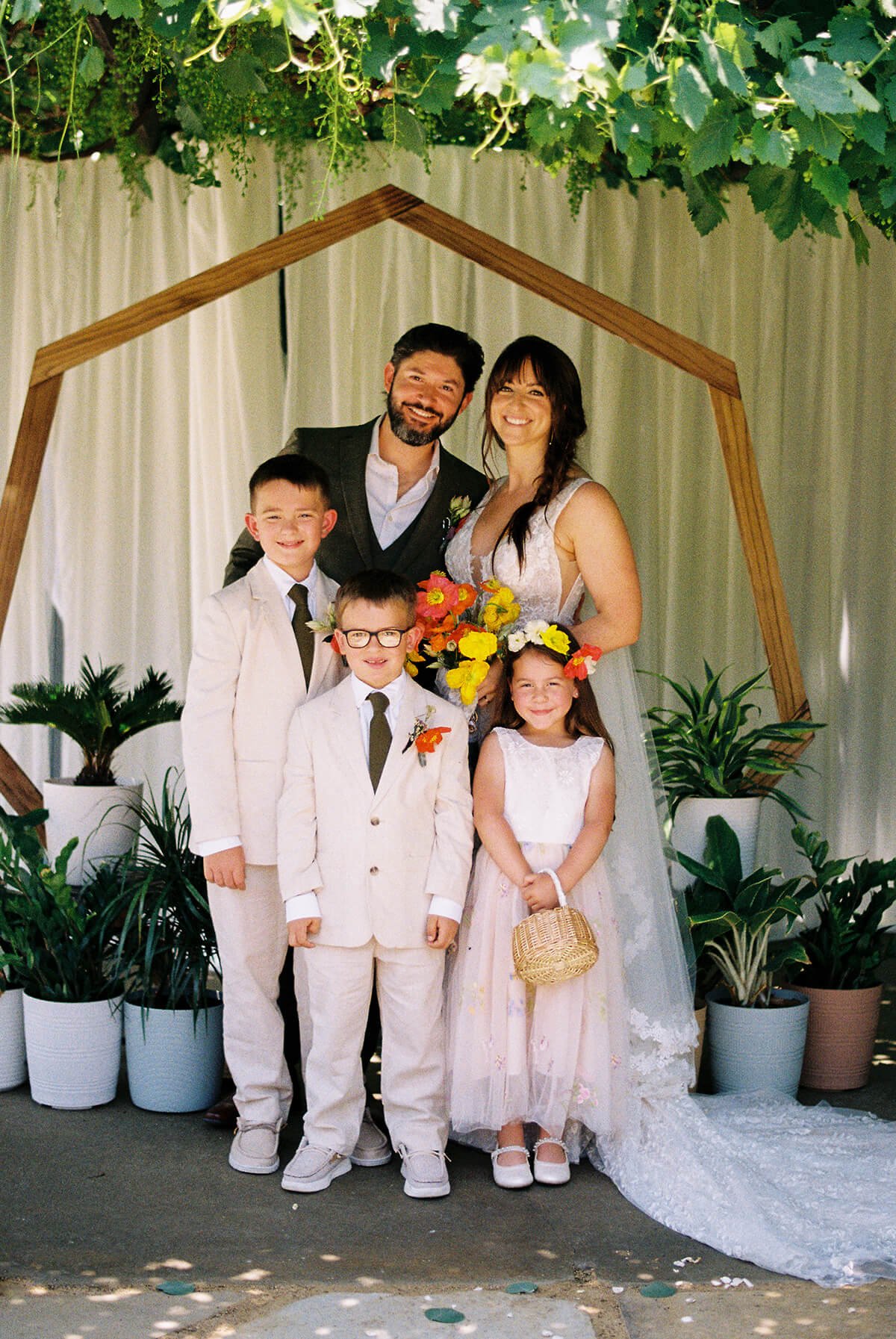 A family at their wedding, standing under a wooden arch decorated with greenery and white flowers, with potted plants around them.