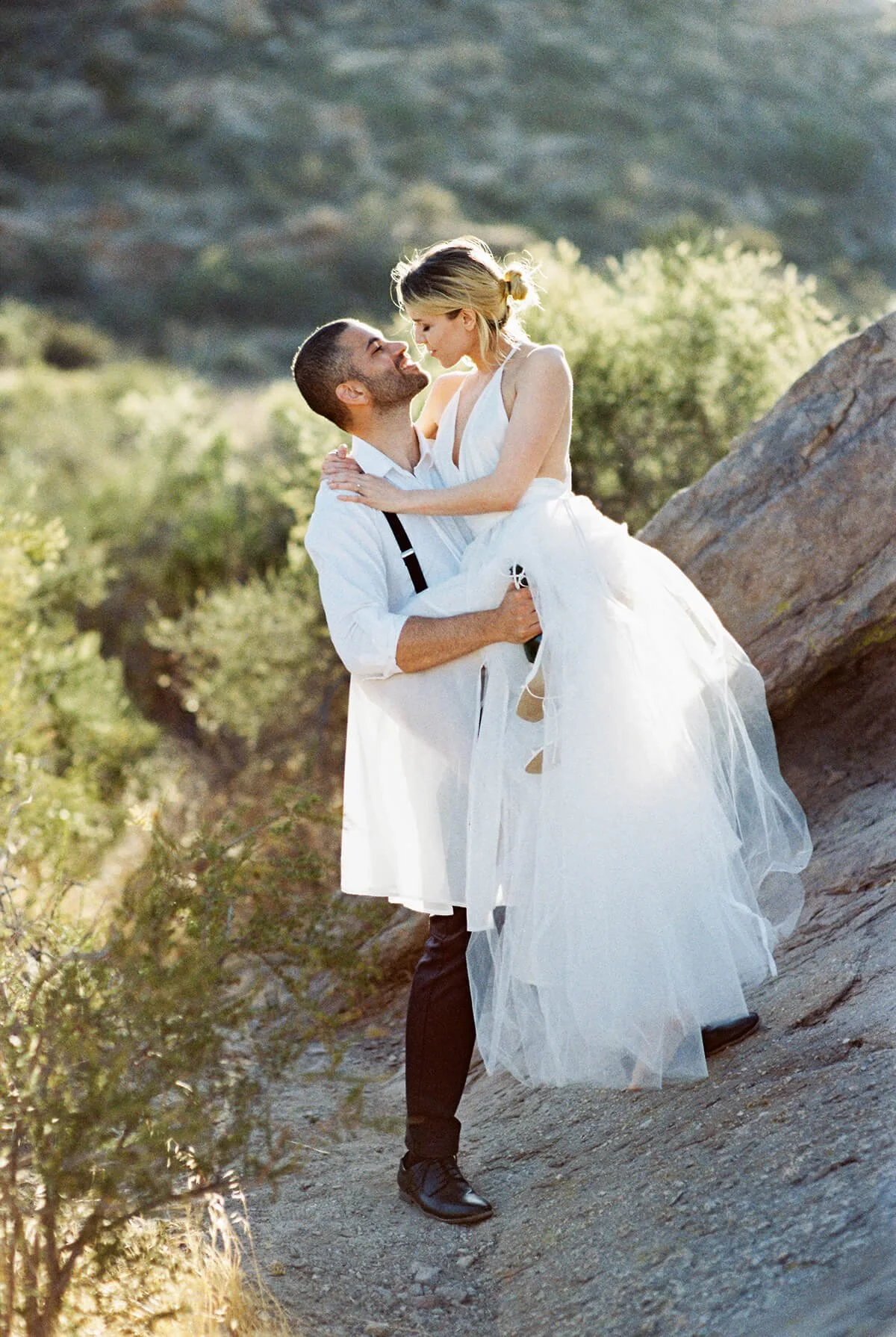 A couple dressed in wedding attire, with the groom in a white shirt and suspenders and the bride in a white wedding gown, standing outdoors on a rocky terrain in Griffith Park with greenery and mountains in the background, sharing an intimate moment.
