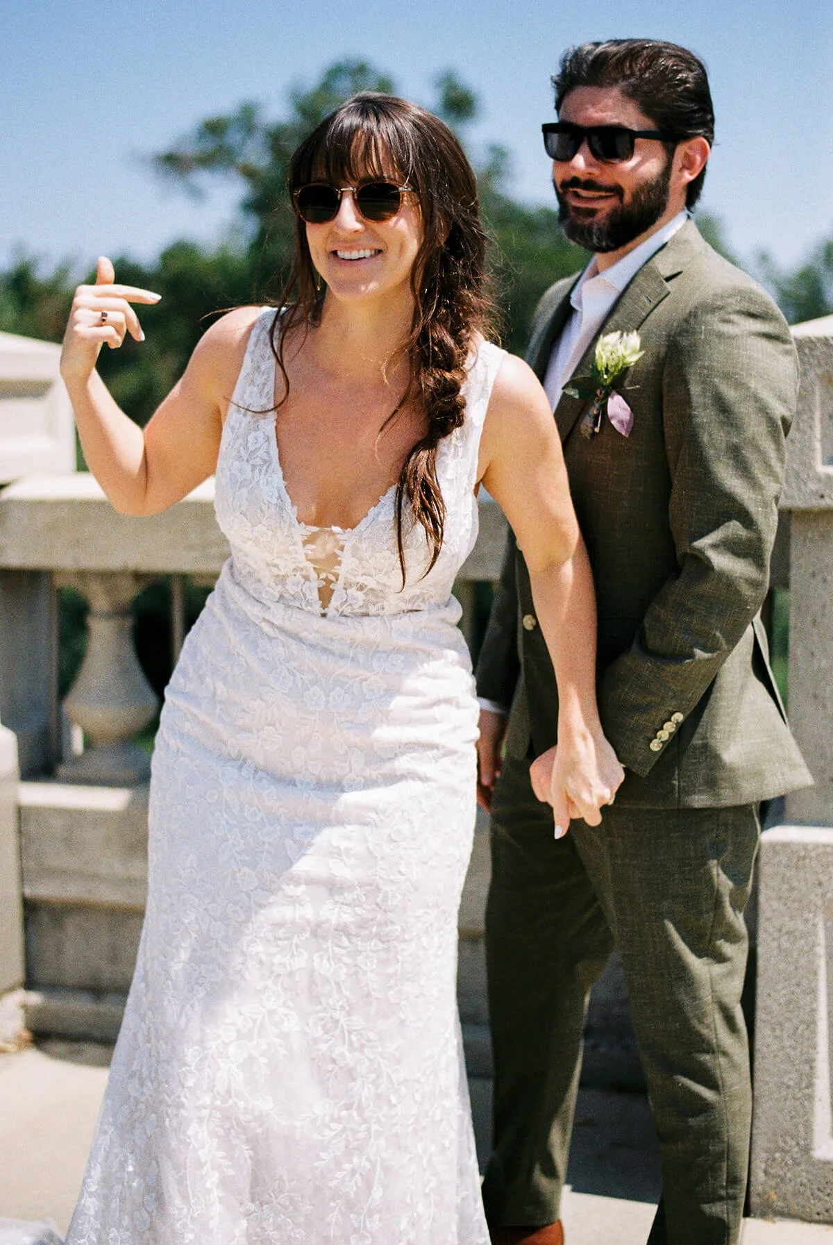 A couple dressed in wedding attire holding hands and smiling outdoors. The woman is wearing a white lace dress and sunglasses, and the man is in a suit with sunglasses, standing in front of a stone railing with a sunny sky and greenery in the backgro