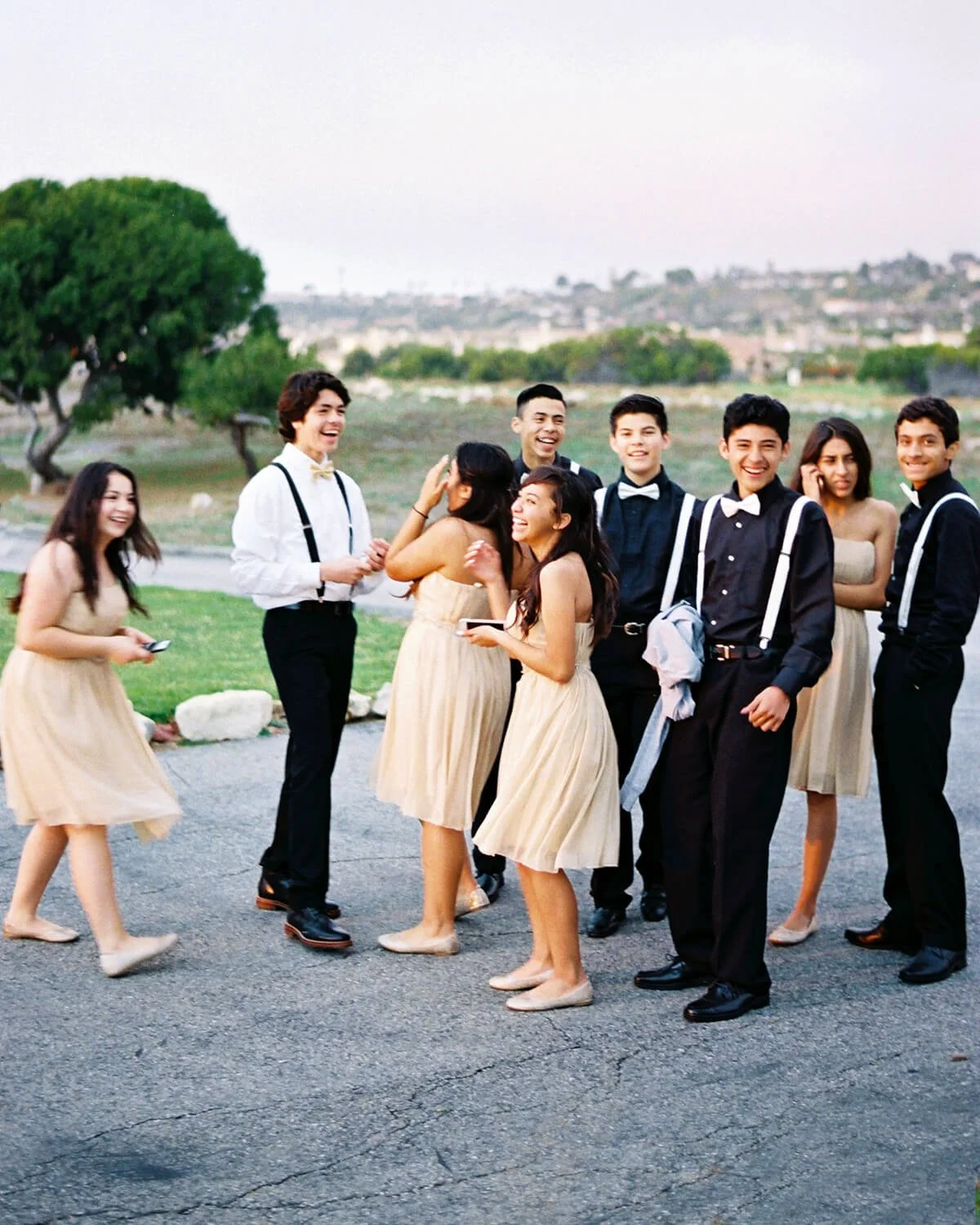 A Quinceañera Court dressed in semi-formal attire, standing outdoors on a paved area, laughing and talking with a park and open landscape in the background.