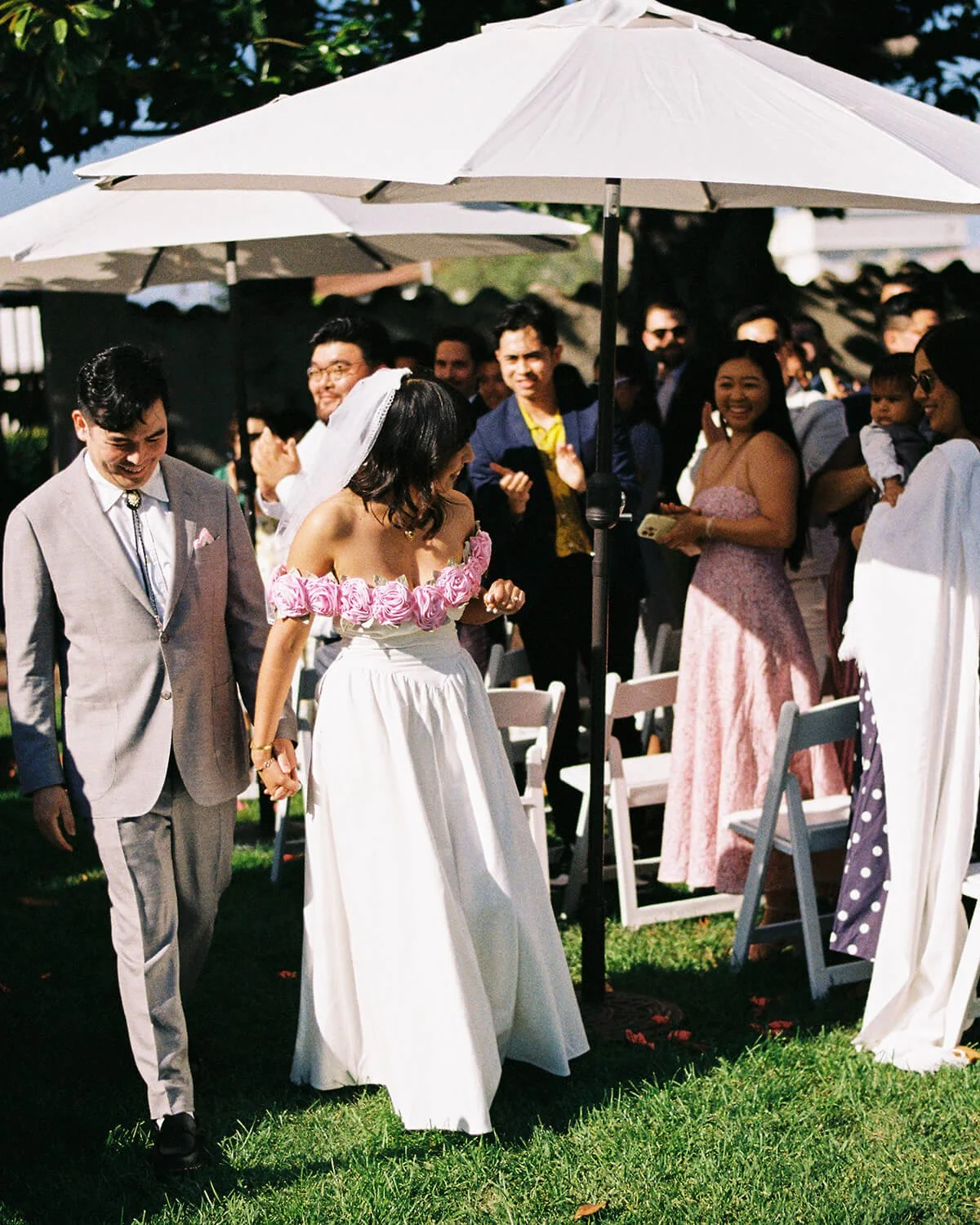 Couple holding hands under white umbrella at outdoor wedding ceremony with guests watching and smiling.